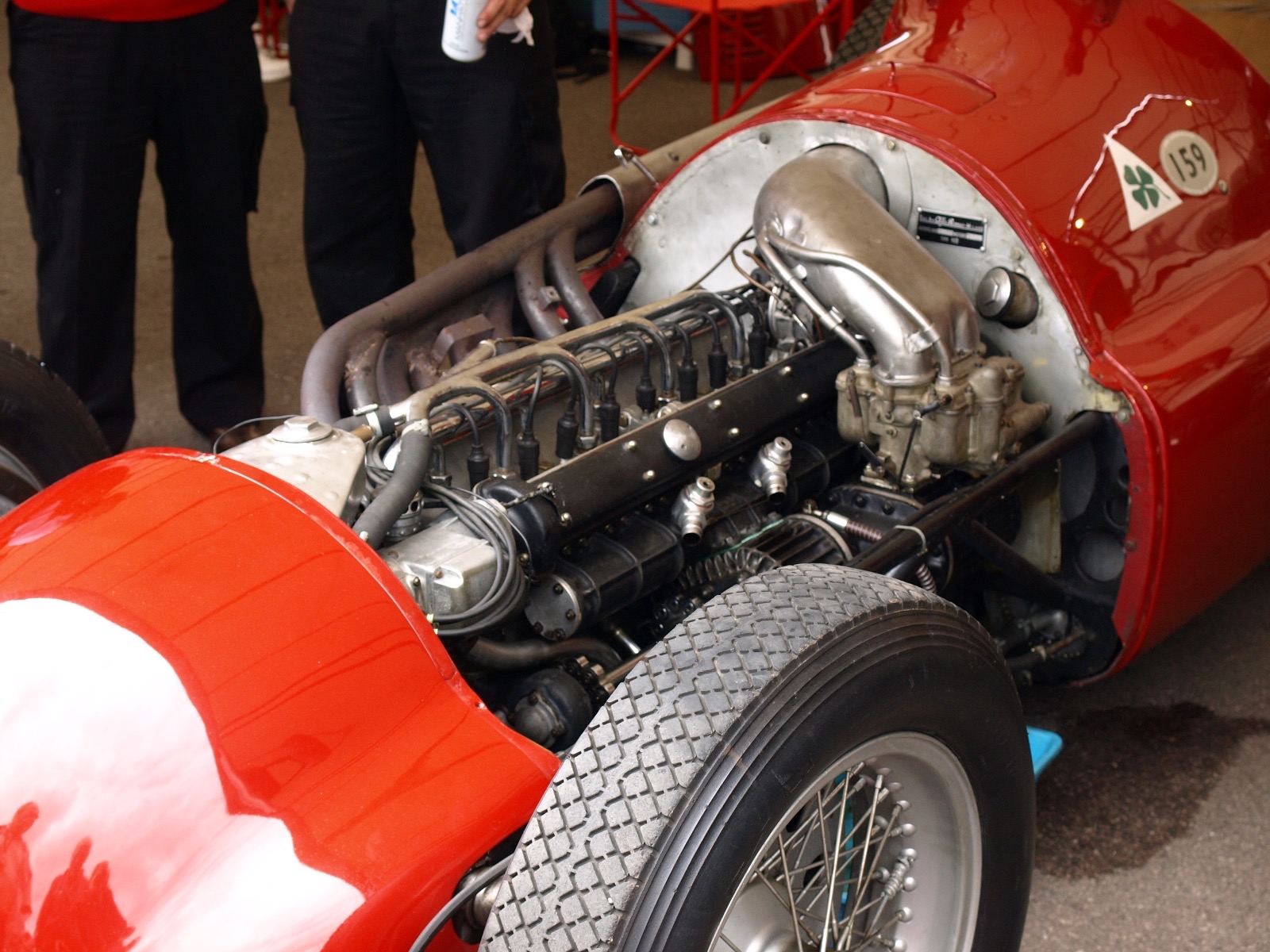 Close view of the straight-eight engine of a red Alfa Romeo 159 with its side panel removed, showing the row of ignition leads, the carburettor and a large curved alloy intake, with a round 159 badge and a green four-leaf clover on the scuttle