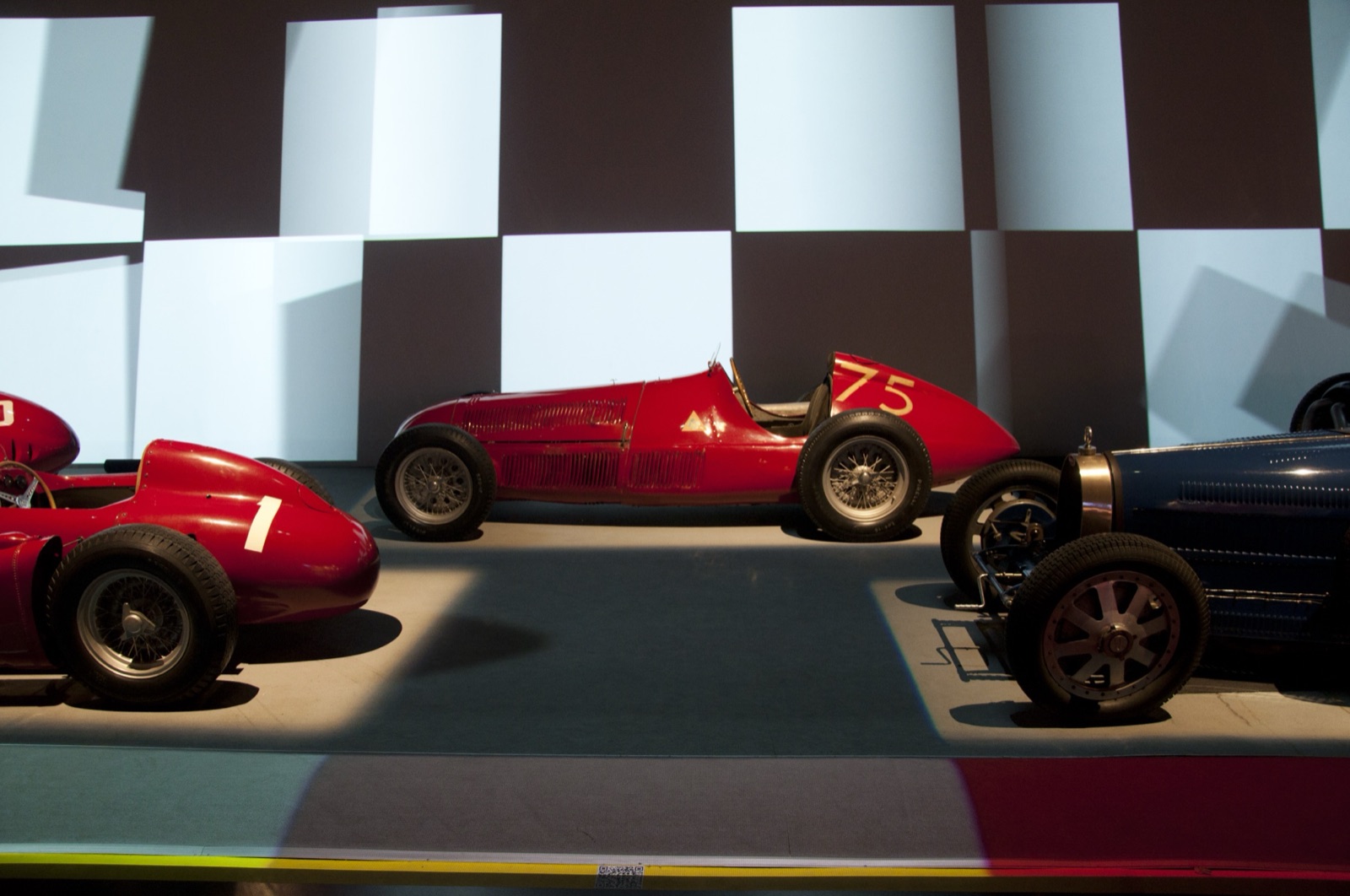 Side view of a red Alfa Romeo 159 single-seater numbered 75 in yellow on its tail, displayed between two other racing cars against a projected chequered pattern