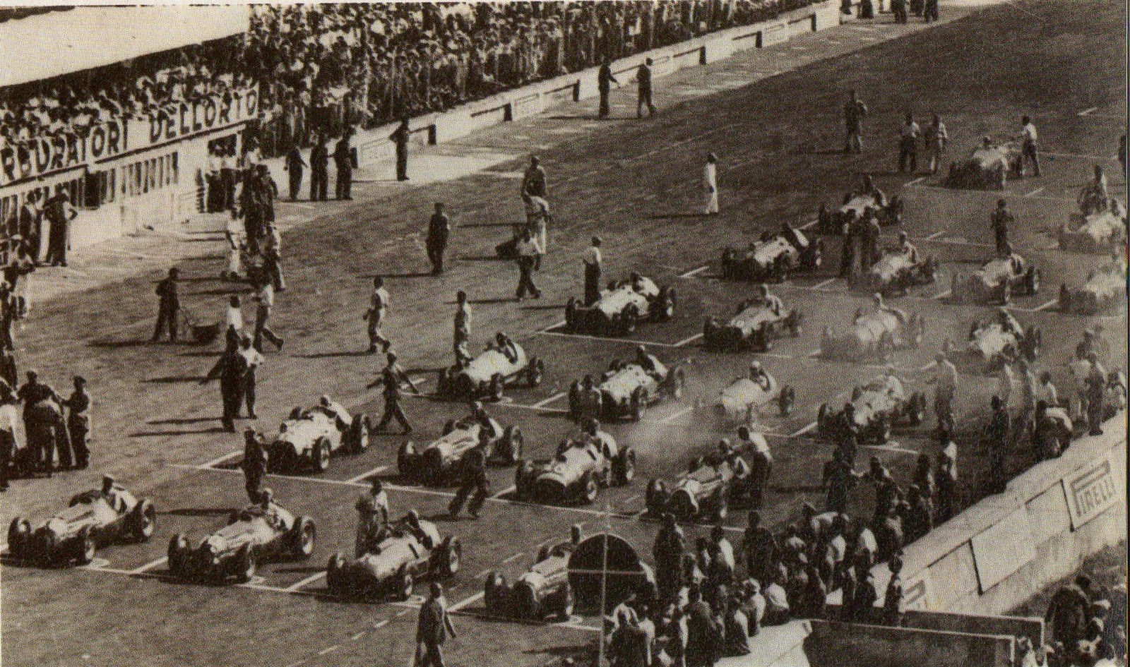 Black and white overhead view of a crowded starting grid at Monza, rows of racing cars with mechanics walking between them, a grandstand and a Dell'Orto banner on the left