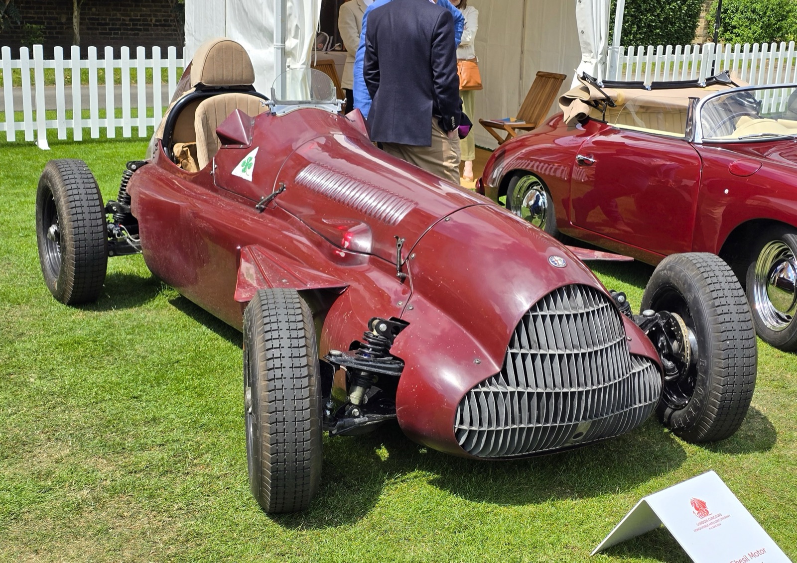 A dark red two-seat car styled on the Alfa Romeo 158, with a slatted grille, exposed coil-spring front suspension and a green four-leaf clover badge, parked on grass beside a red 356-style cabriolet