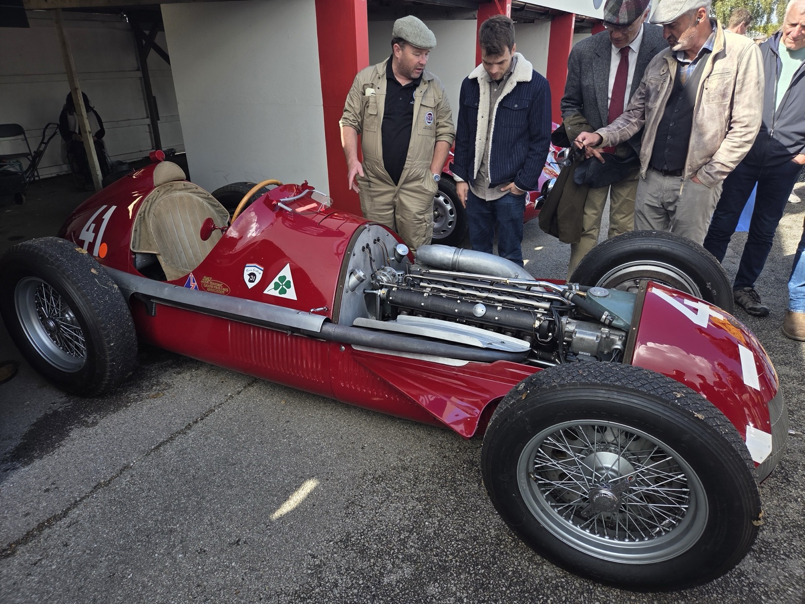 A dark red Alfa Romeo 158 numbered 41 with its bonnet panels off, showing the long straight-eight engine with a long alloy pipe above it and the exhaust manifold below, in a paddock garage