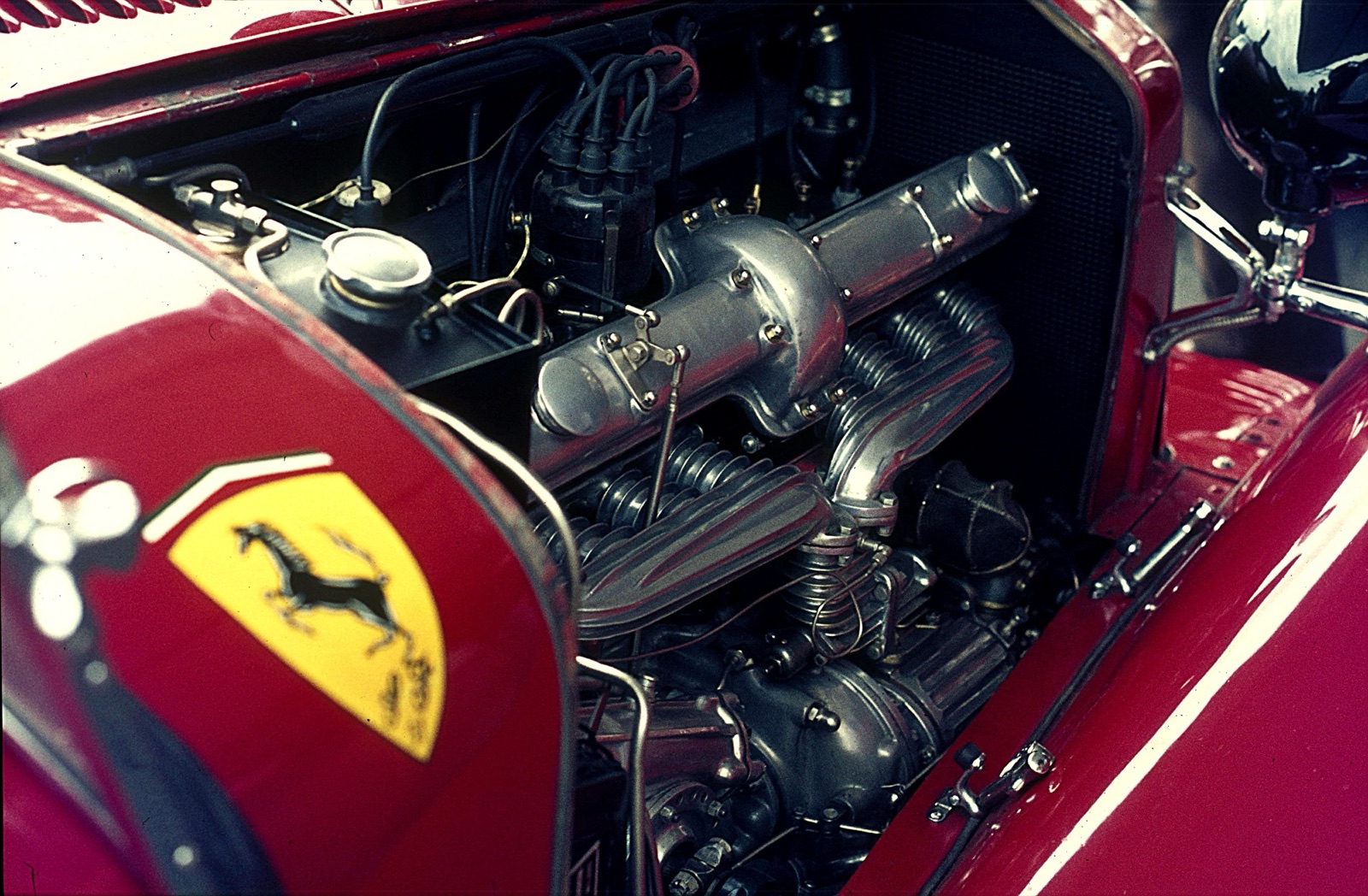 The engine bay of a red Alfa Romeo with a Scuderia Ferrari shield on the bonnet side, showing polished alloy castings and ribbed pipes along the straight-eight