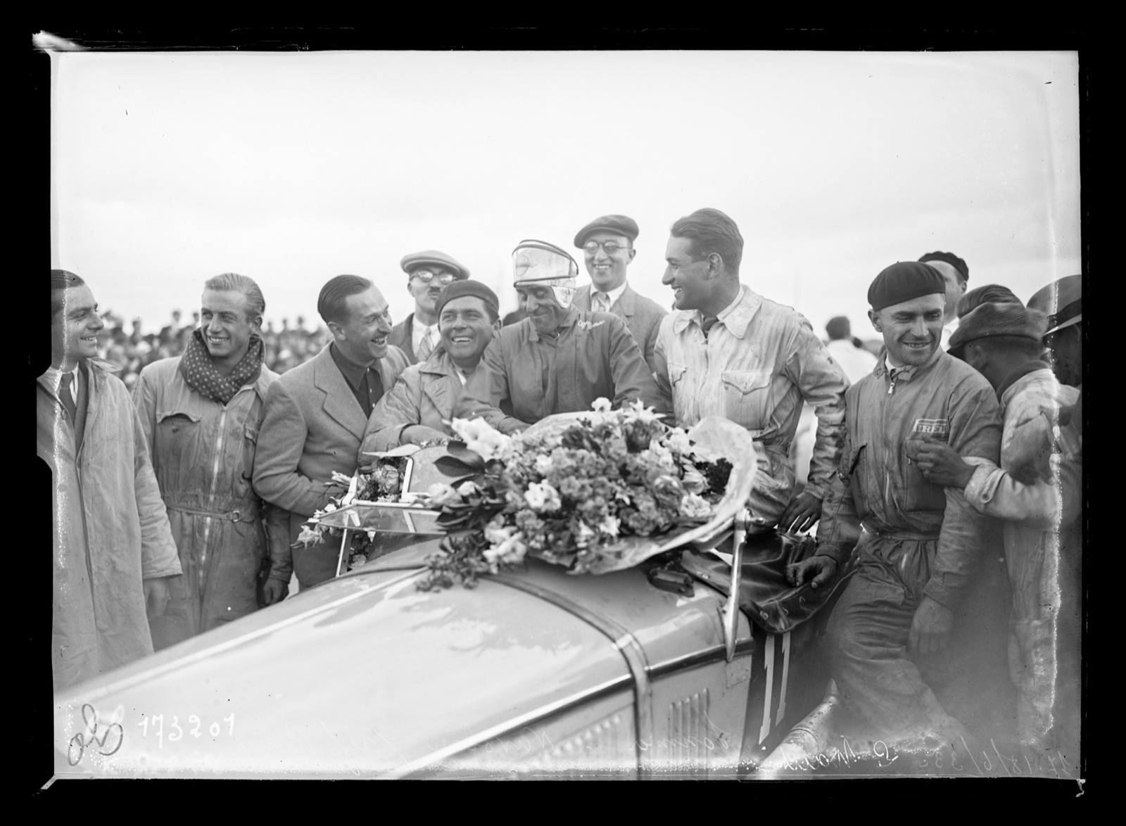 Smiling men in overalls, caps and berets crowd round the bonnet of a racing car laden with a large bouquet of flowers
