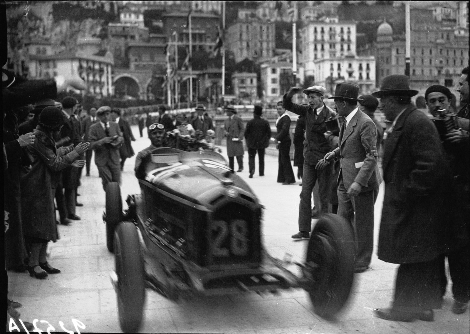 A racing car numbered 28 driven slowly among spectators in coats and hats on a Monte Carlo street, buildings on the hillside behind