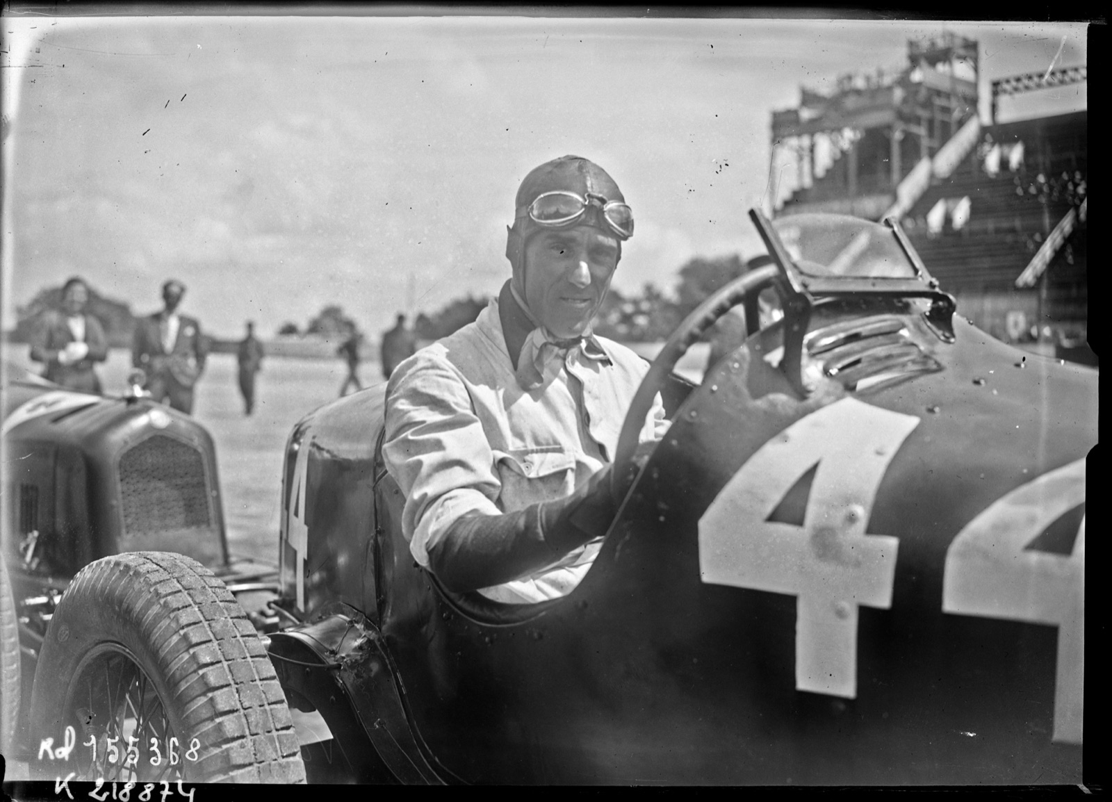 Tazio Nuvolari in a leather helmet with goggles on his forehead, seated in a dark racing car with the number 44 on its side, a grandstand behind