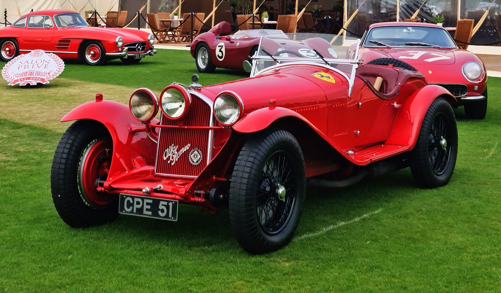 A red Alfa Romeo 8C 2300 Monza with cycle wings, two headlamps and a central lamp, parked on a lawn with other red cars behind