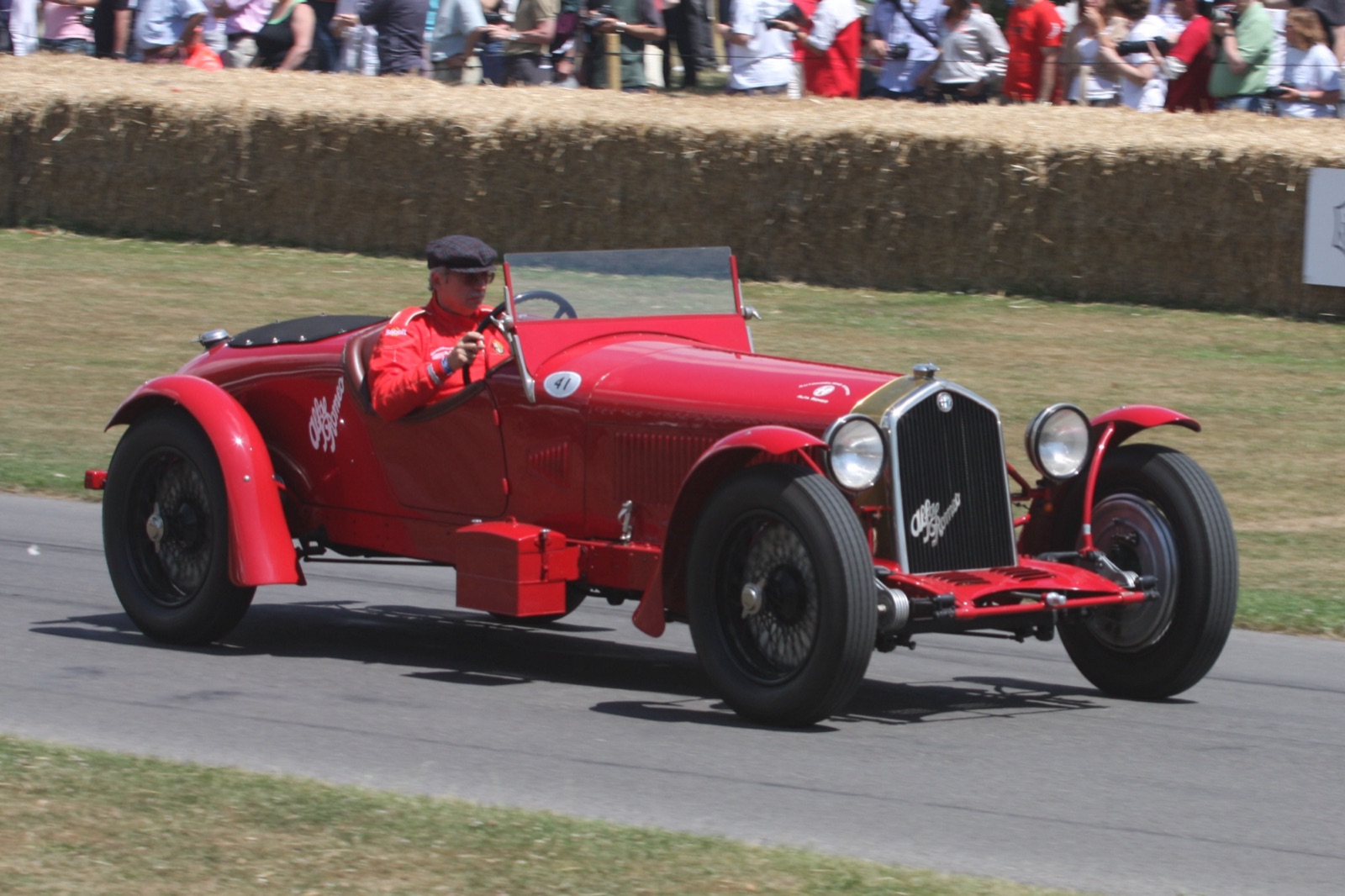 A red long-chassis Alfa Romeo sports car with flowing wings and a box on its running board, driven past straw bales and spectators