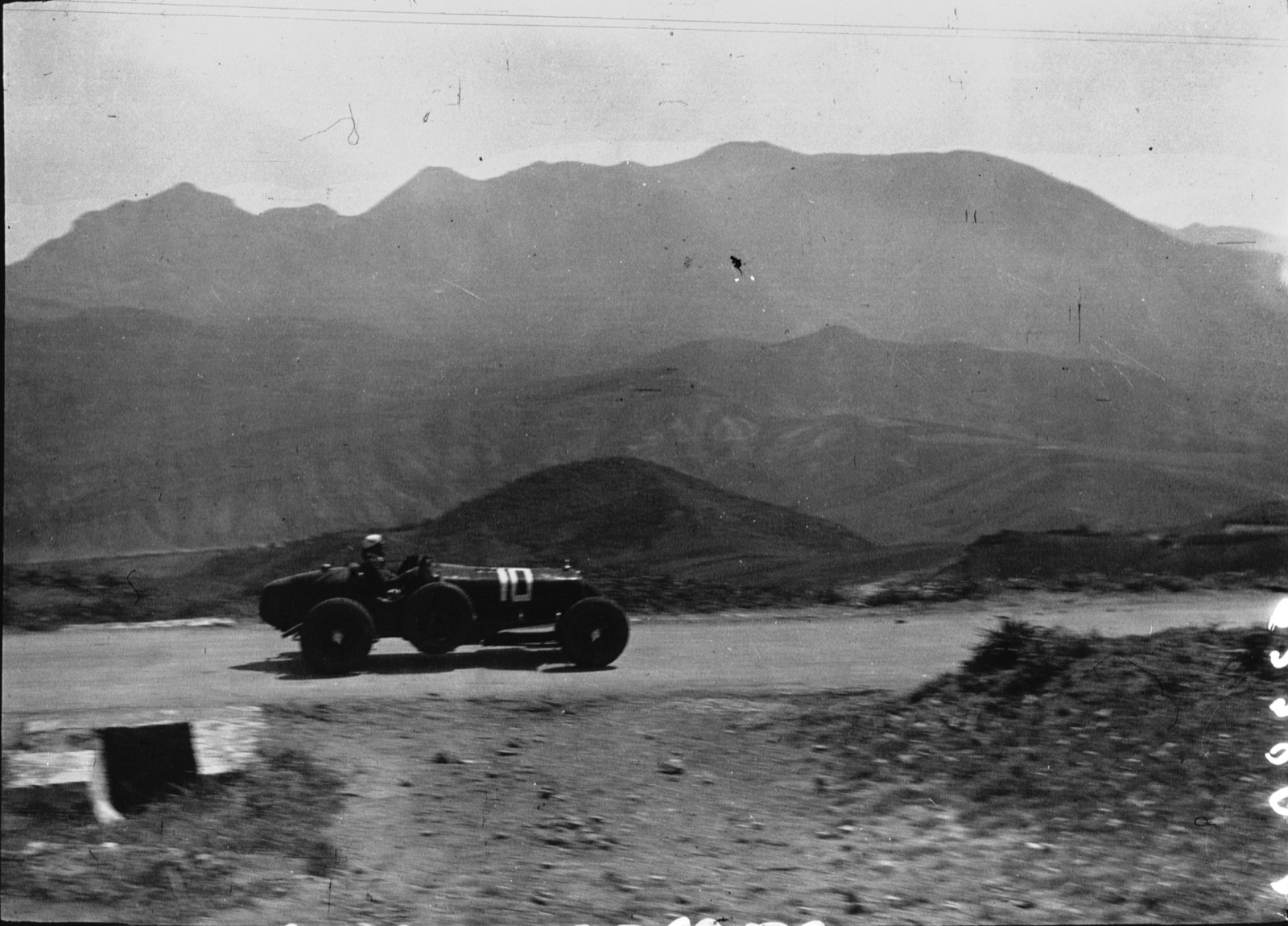 A dark open racing car numbered 10 on a dusty mountain road, bare mountains behind