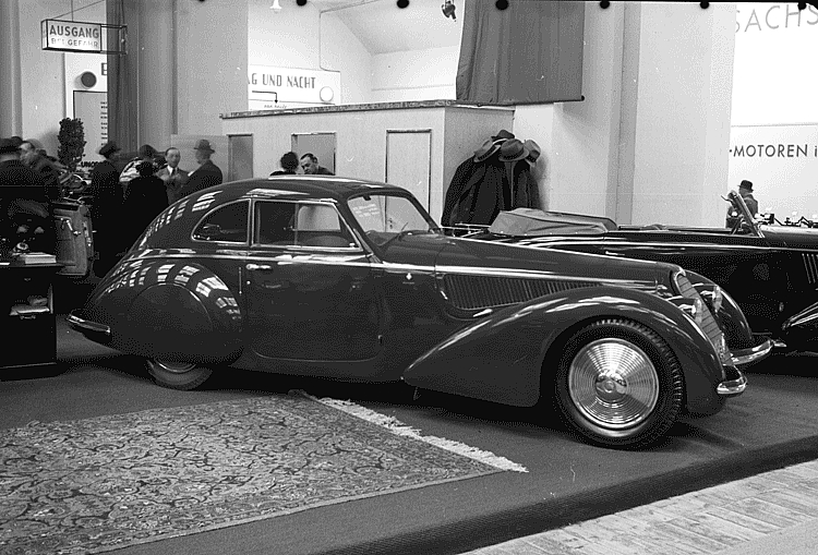Black and white photograph of a dark fastback coupé with enclosed rear wheels and a louvred bonnet on a motor show stand, men in hats behind