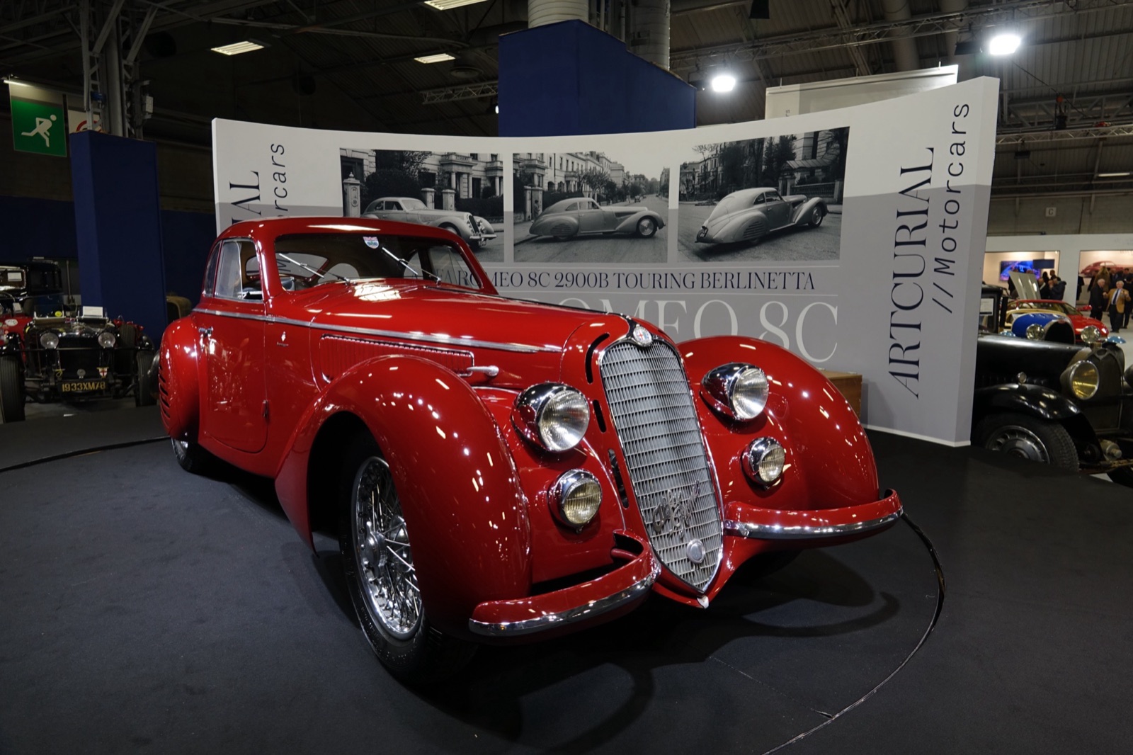 A red Touring fastback coupé with chrome bumpers and a tall grille on an auction stand, a banner reading Alfa Romeo 8C 2900B Touring Berlinetta behind it