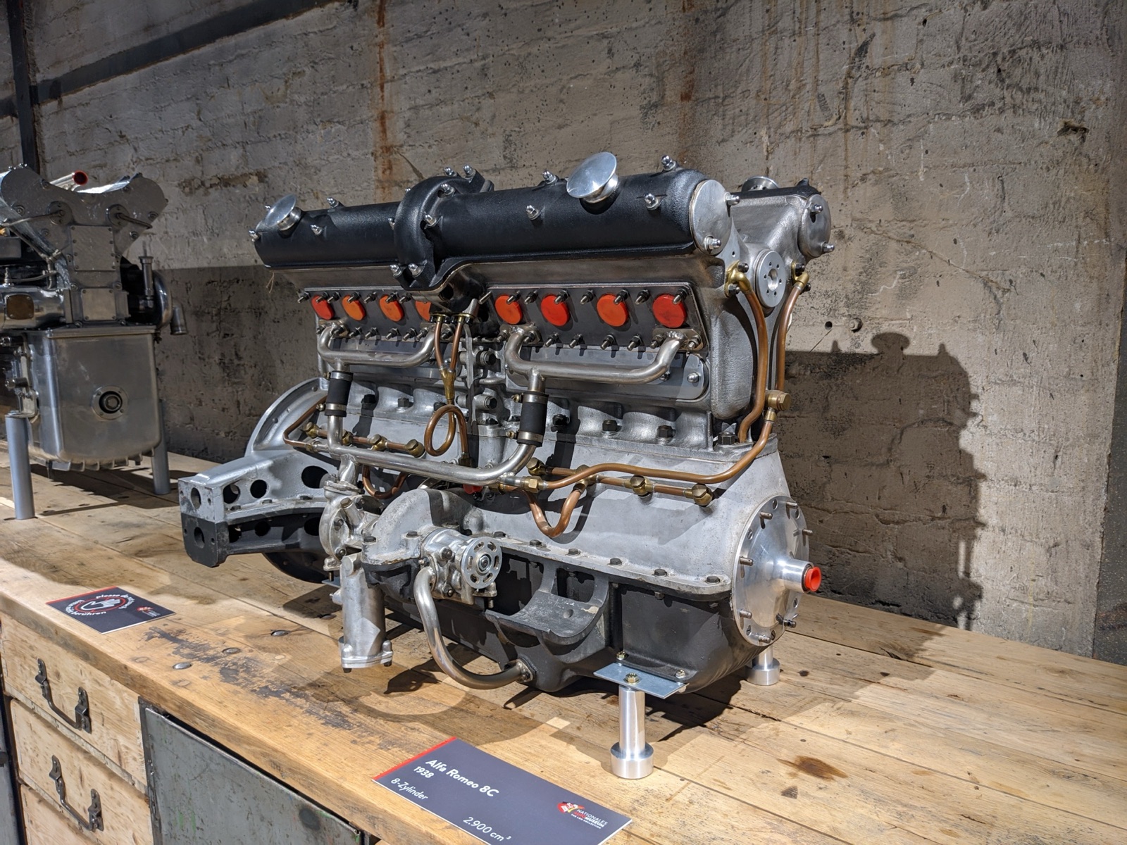 A bare straight-eight engine with two black camshaft covers, eight red plug caps and copper pipework, on a wooden bench against a concrete wall