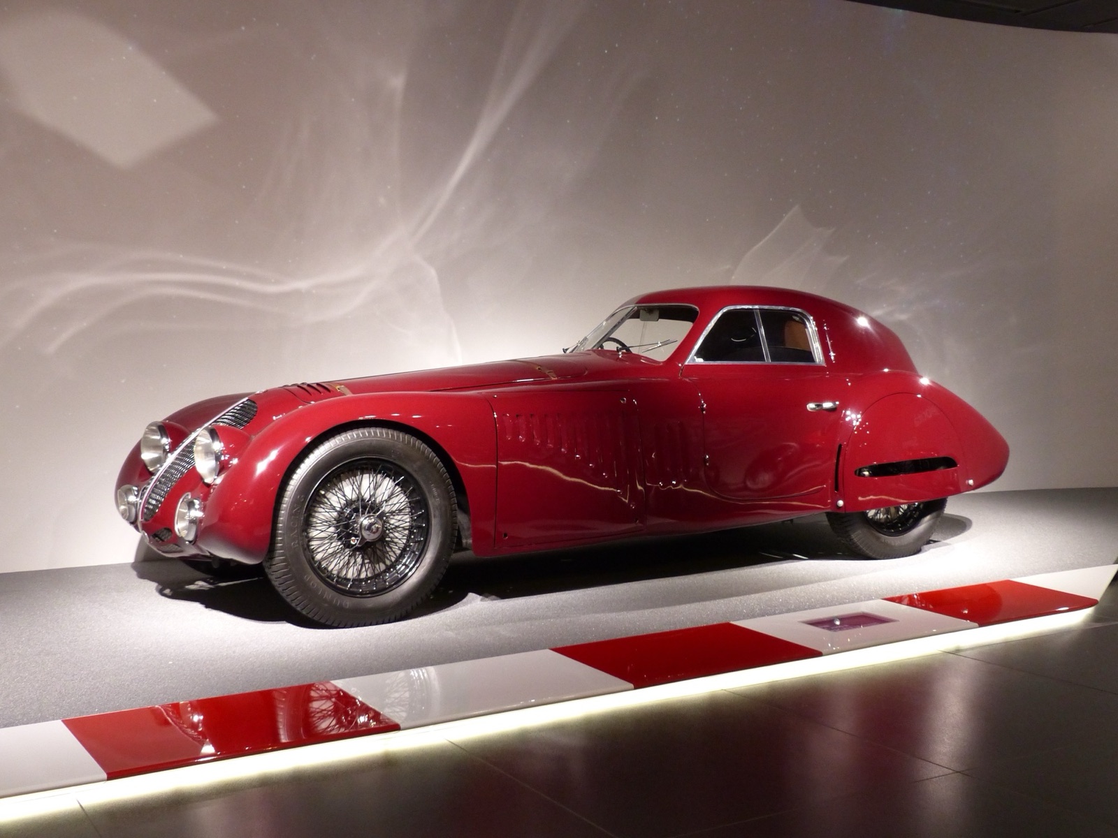 A dark red streamlined fastback coupé with enclosed rear wheels, louvred sides and wire wheels, on a lit museum plinth