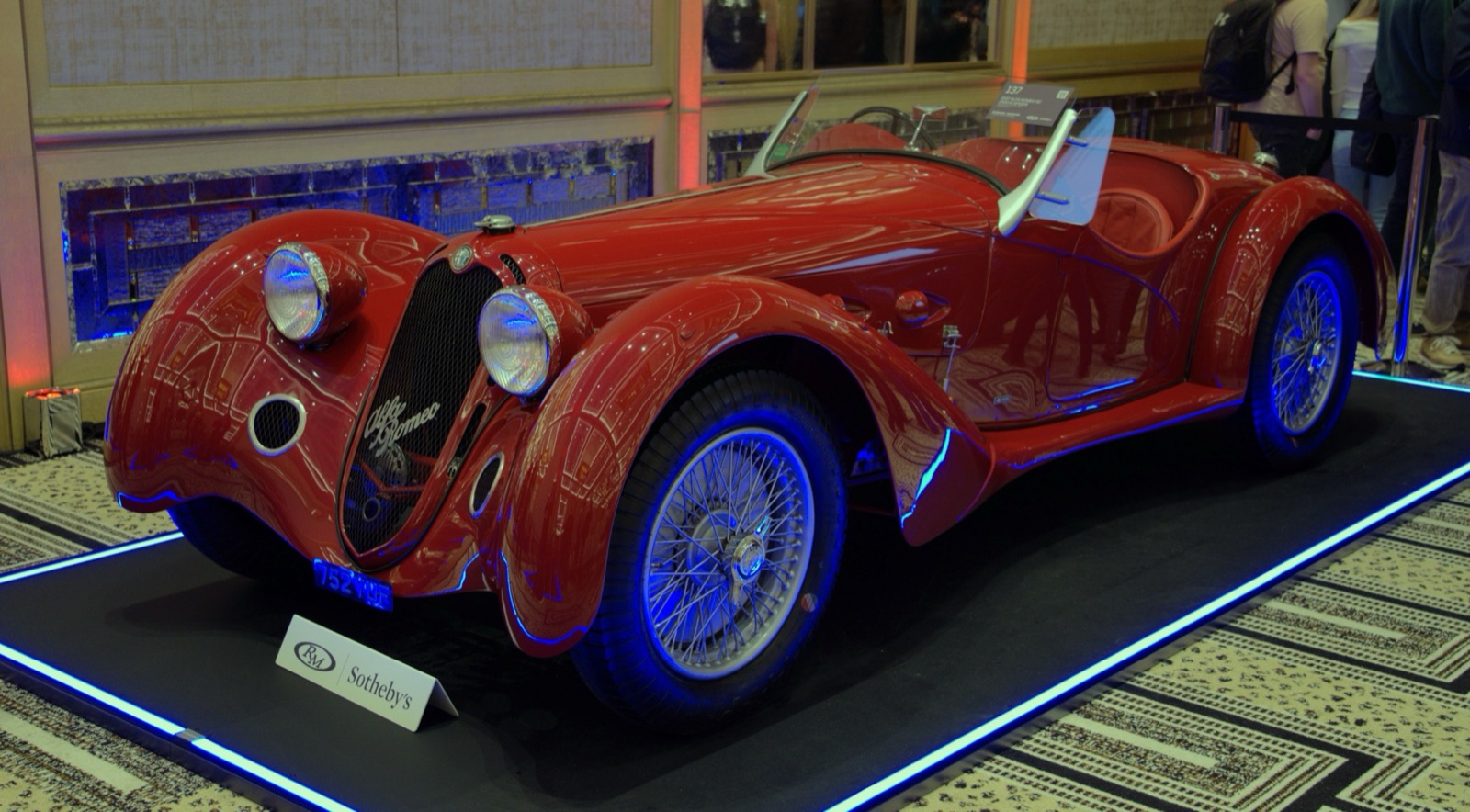 A red open two-seater with swept wings, wire wheels and an Alfa Romeo script on the grille, under blue lights on an auction display