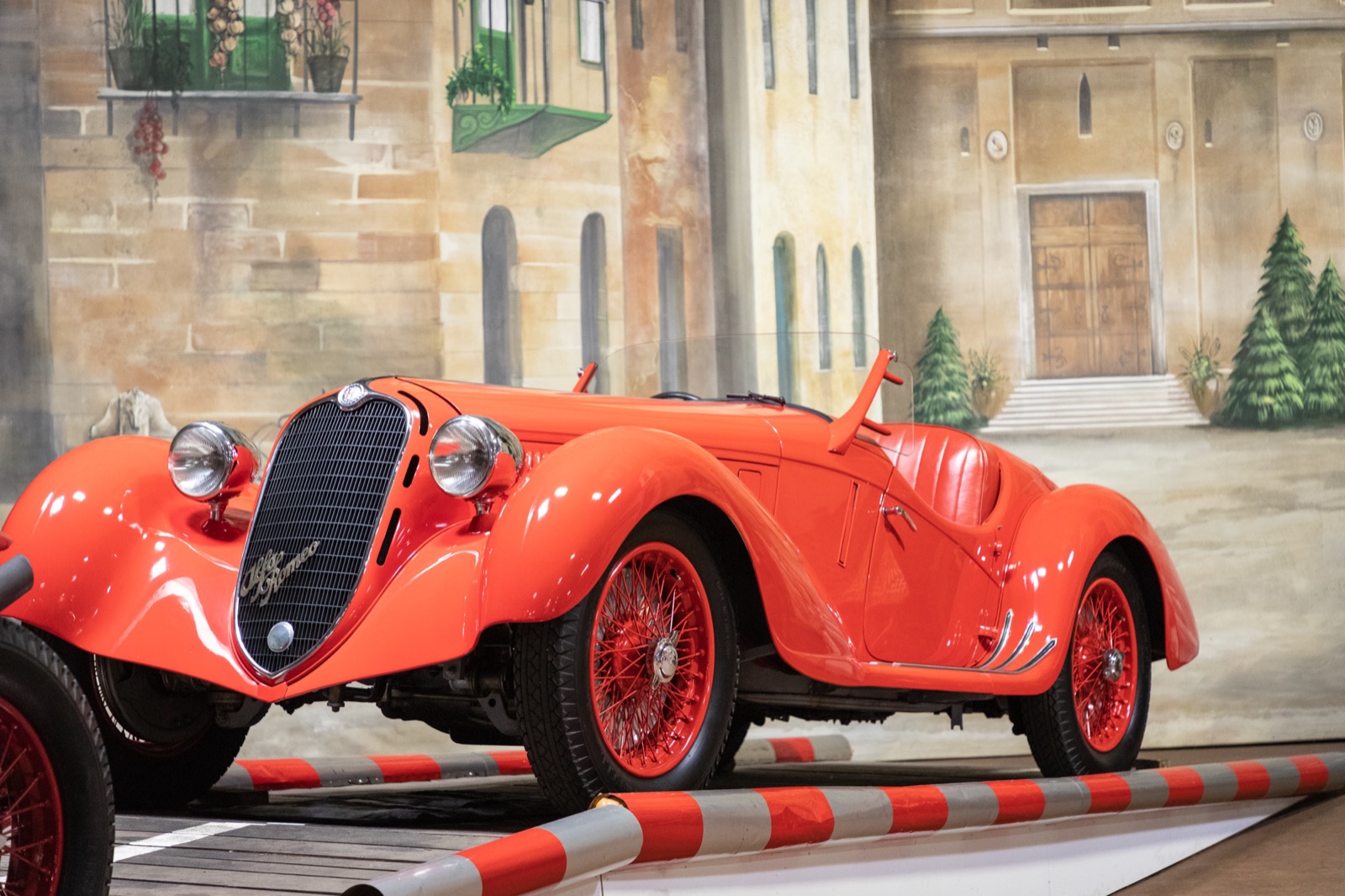 A red open two-seat Alfa Romeo with flowing wings, red wire wheels and an upright grille, on a museum stand painted as an Italian street
