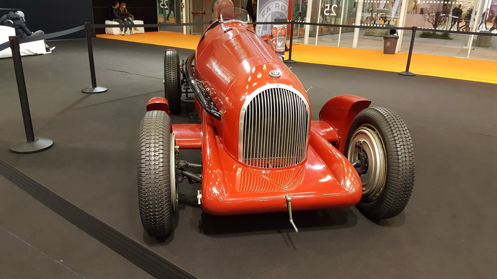 Front view of a red Alfa Romeo single-seater with a fully enclosed streamlined body, a large chrome grille and faired front wheels, on show behind a rope barrier