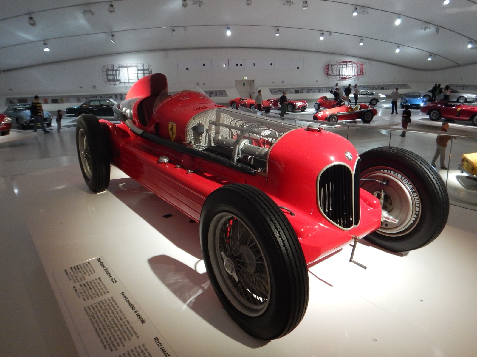 A red Alfa Romeo Bimotore with a long bonnet, a second engine visible under a mesh cover behind the cockpit, on display in a white museum hall