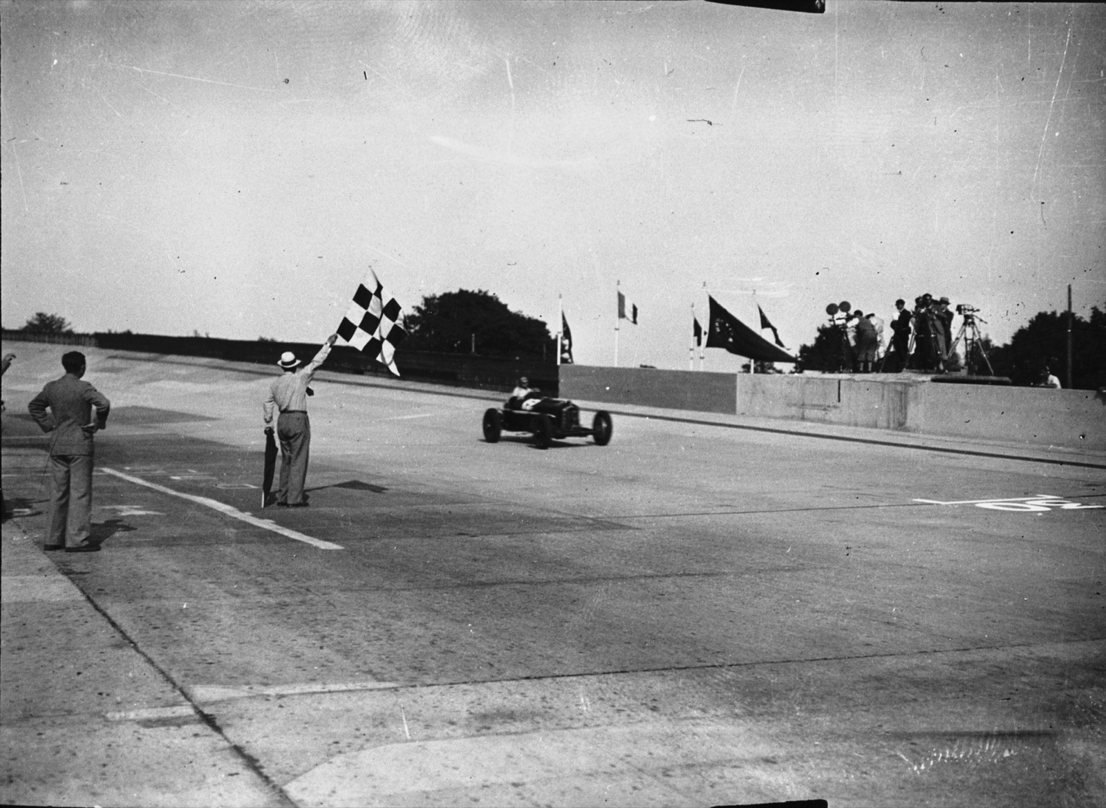 A starter waving a chequered flag as a single dark racing car passes the finish line on a wide concrete track, with flags and film cameras on a stand behind