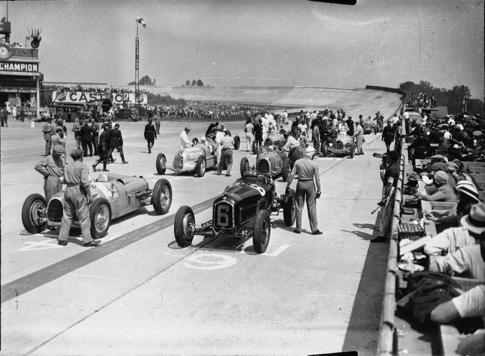 Racing cars lined up on the concrete start area at Montlhéry: a dark Alfa Romeo numbered 6 in the foreground beside a silver German car, with more silver German cars and the banked track behind