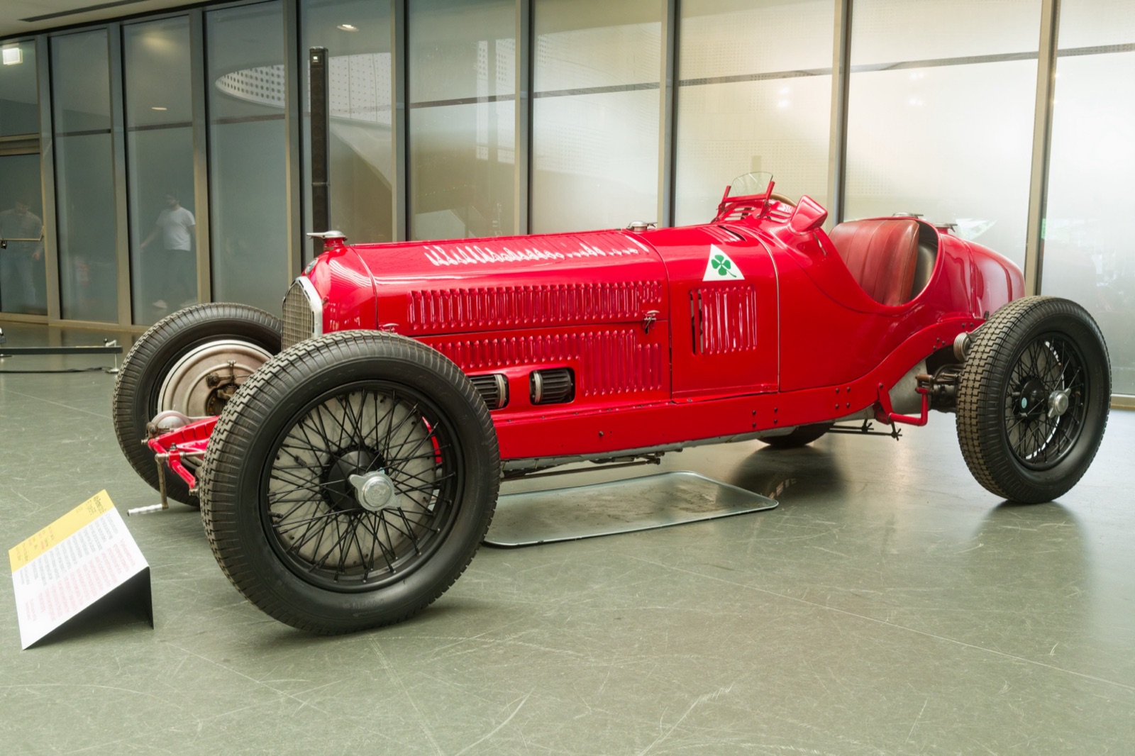A red Alfa Romeo P3 single-seater with a narrow body, wire wheels and a green four-leaf clover badge, on display in a museum hall
