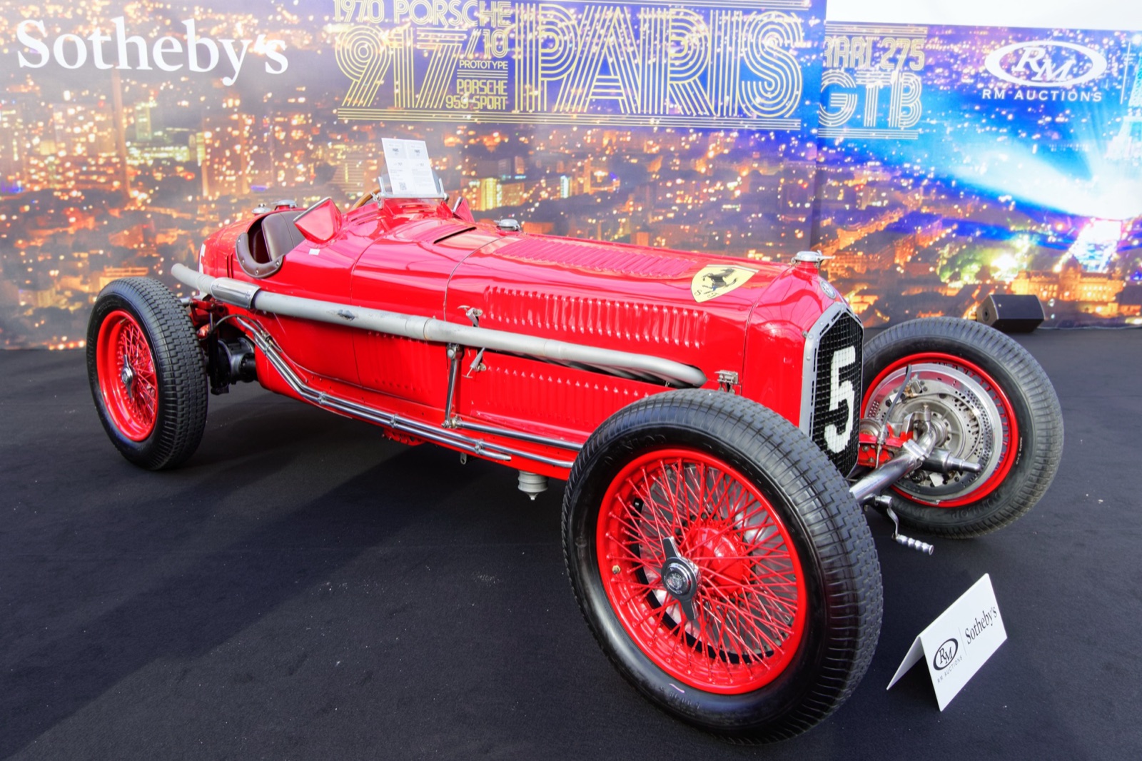 A red Alfa Romeo P3 numbered 5 with red wire wheels and independent front suspension, displayed on a black carpet in front of a backdrop printed with car names