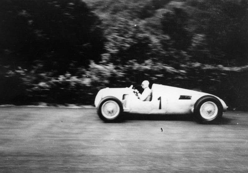 Bernd Rosemeyer at speed in a white Auto Union Type C with the number 1 on its flank, trees blurred behind