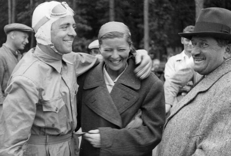Bernd Rosemeyer in racing overalls with his arm round Elly Beinhorn, beside a smiling Ferdinand Porsche in a felt hat