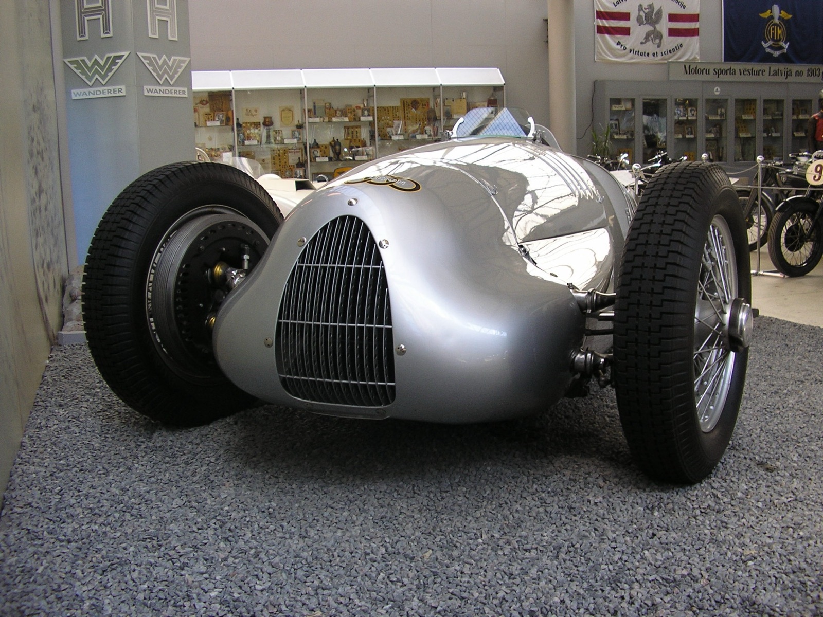 A polished silver Auto Union Type C/D hill-climb car on display on a gravel bed in a museum hall