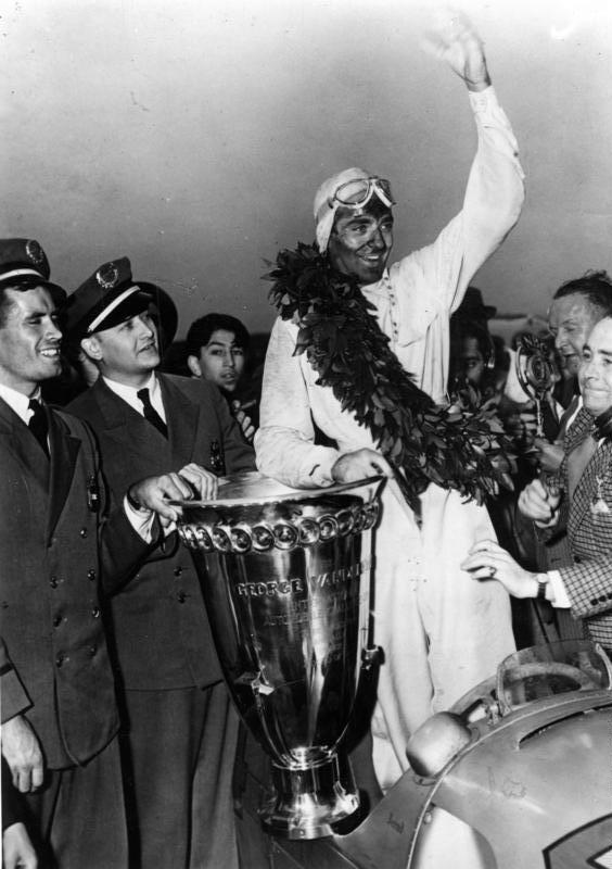 Bernd Rosemeyer in a laurel wreath, waving from the cockpit beside the large Vanderbilt Cup trophy, surrounded by officials