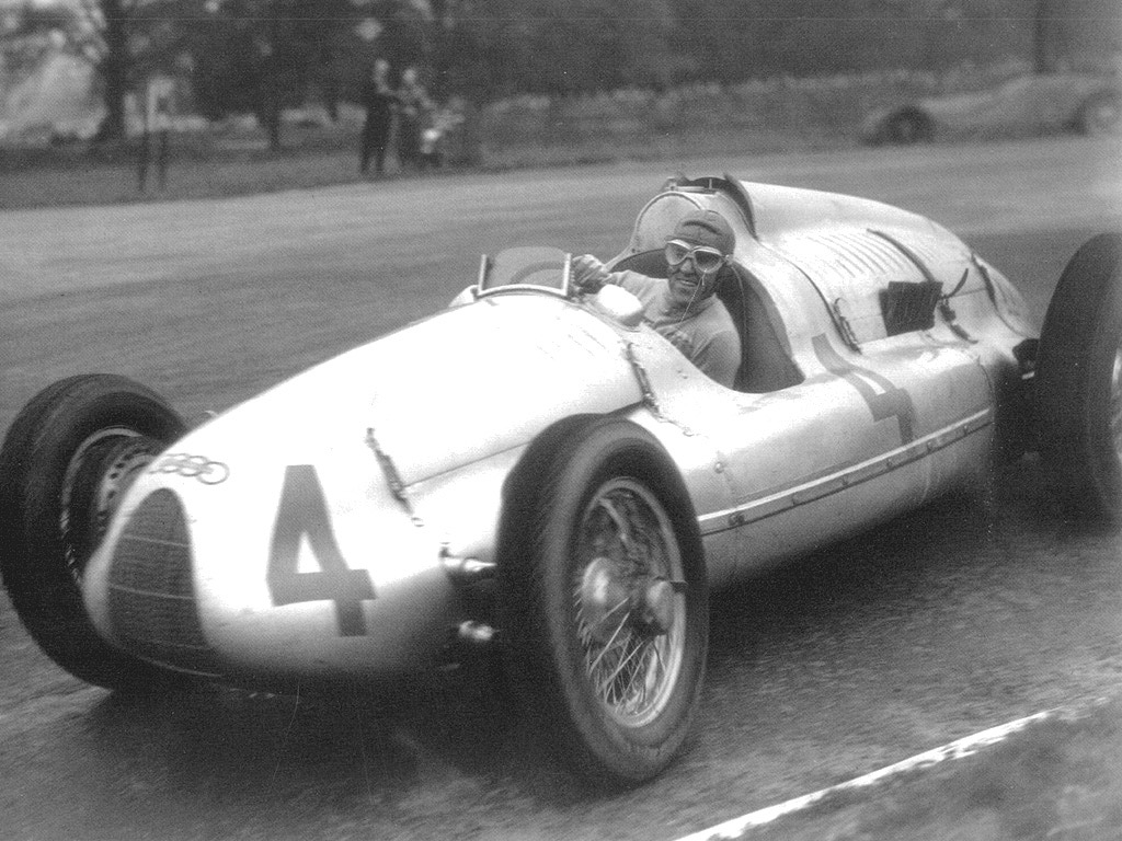 A black-and-white photograph of a smiling driver in goggles and a cap at the wheel of a silver racing car numbered 4, cornering on a street
