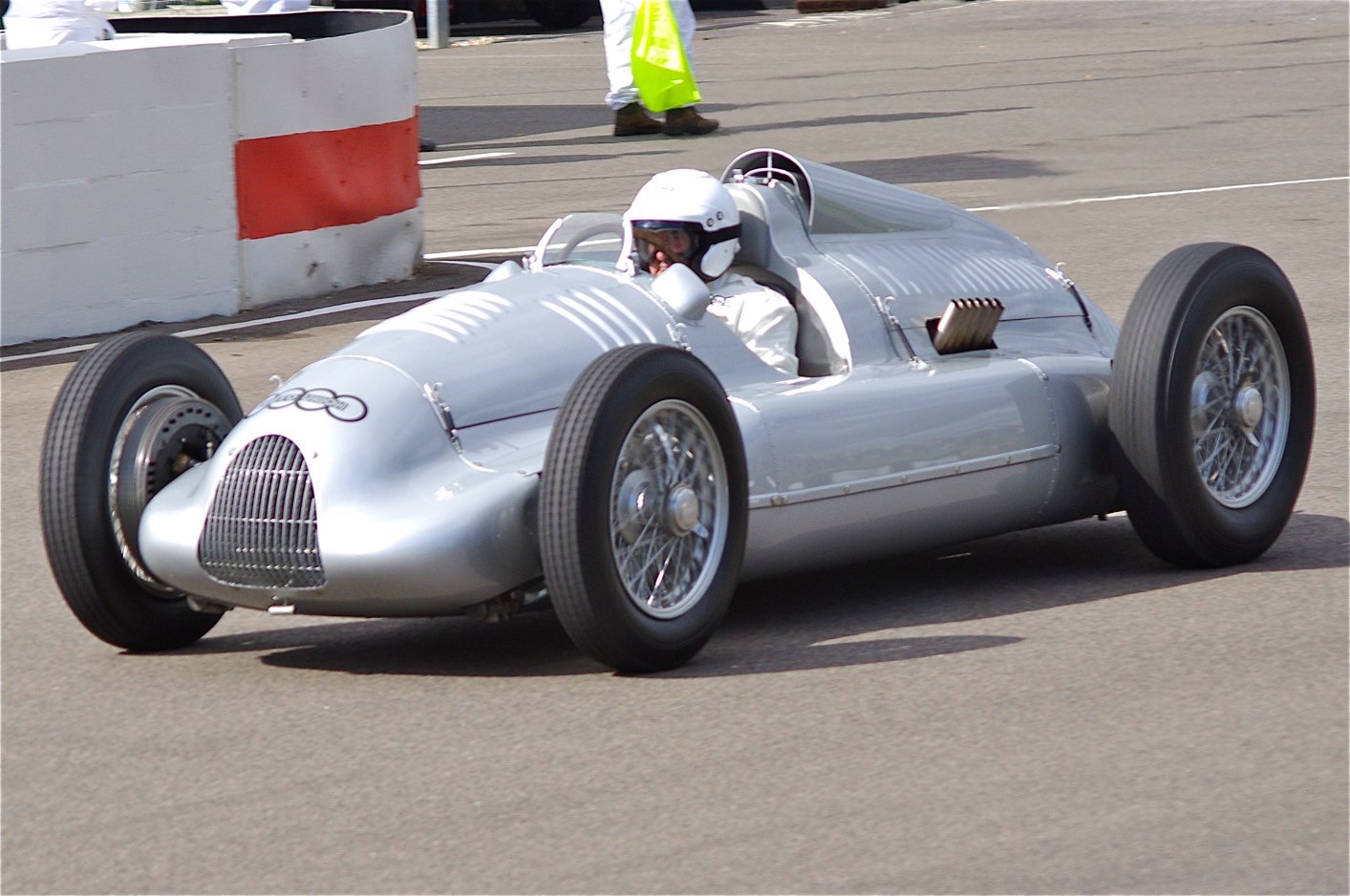 A silver single-seat racing car with a grilled nose and wire wheels turning on a track, its driver in a white helmet, beside a red and white barrier