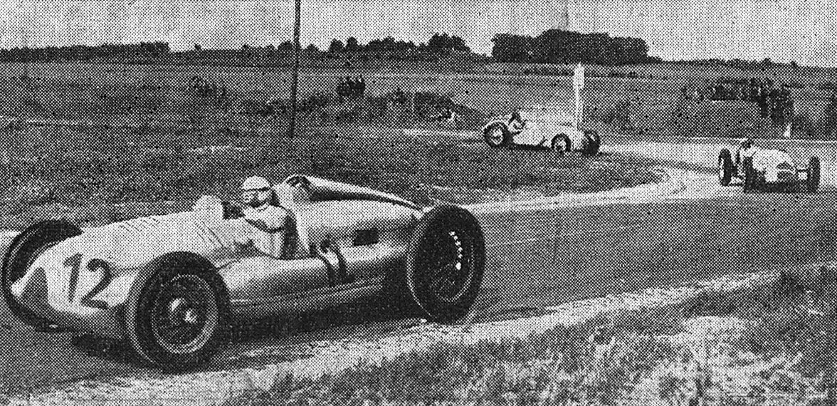 A newspaper photograph of a silver racing car numbered 12 rounding a corner on a country road, with two cars following in the distance