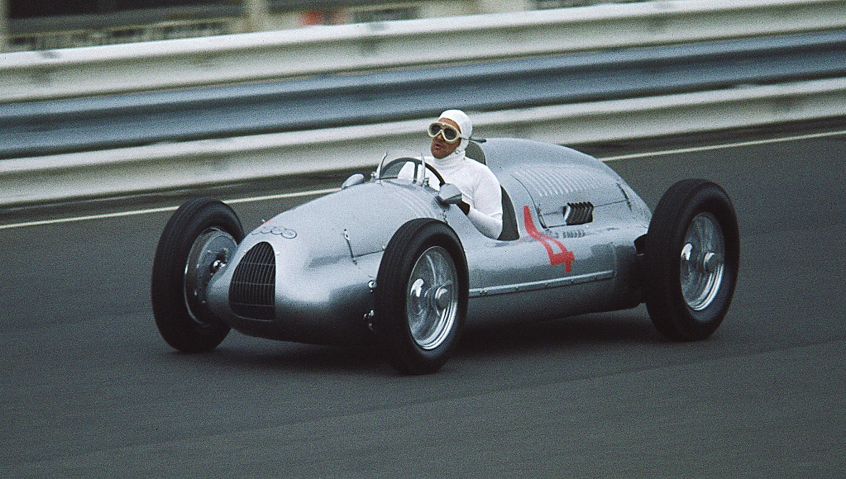 A silver racing car numbered 4 on a wide track with Armco barriers, its driver in a white helmet cover and goggles