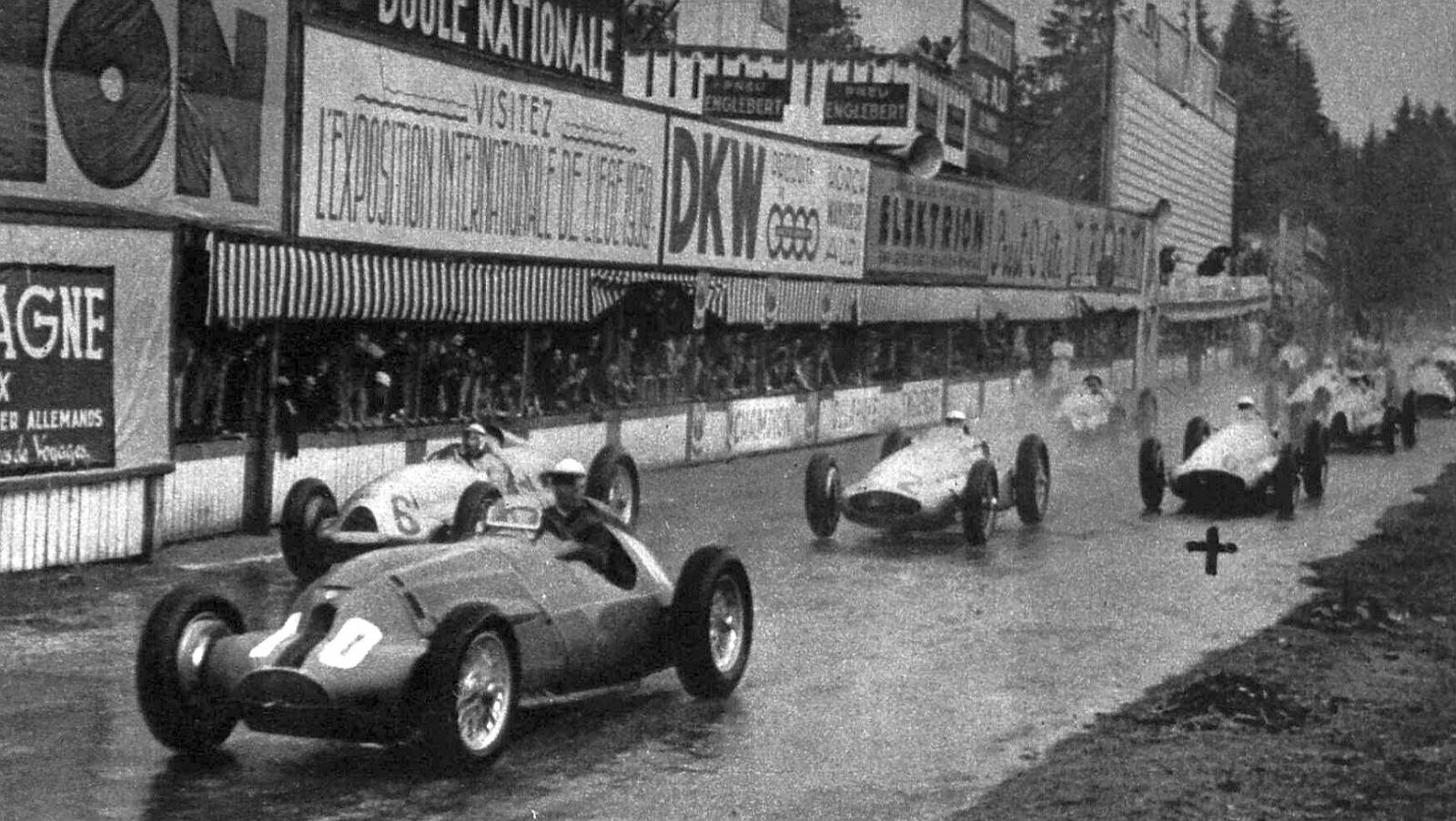A wet start-line straight lined with advertising hoardings and a covered stand, with a dark Alfa Romeo numbered 10 leading a silver Auto Union numbered 6 and two other silver cars