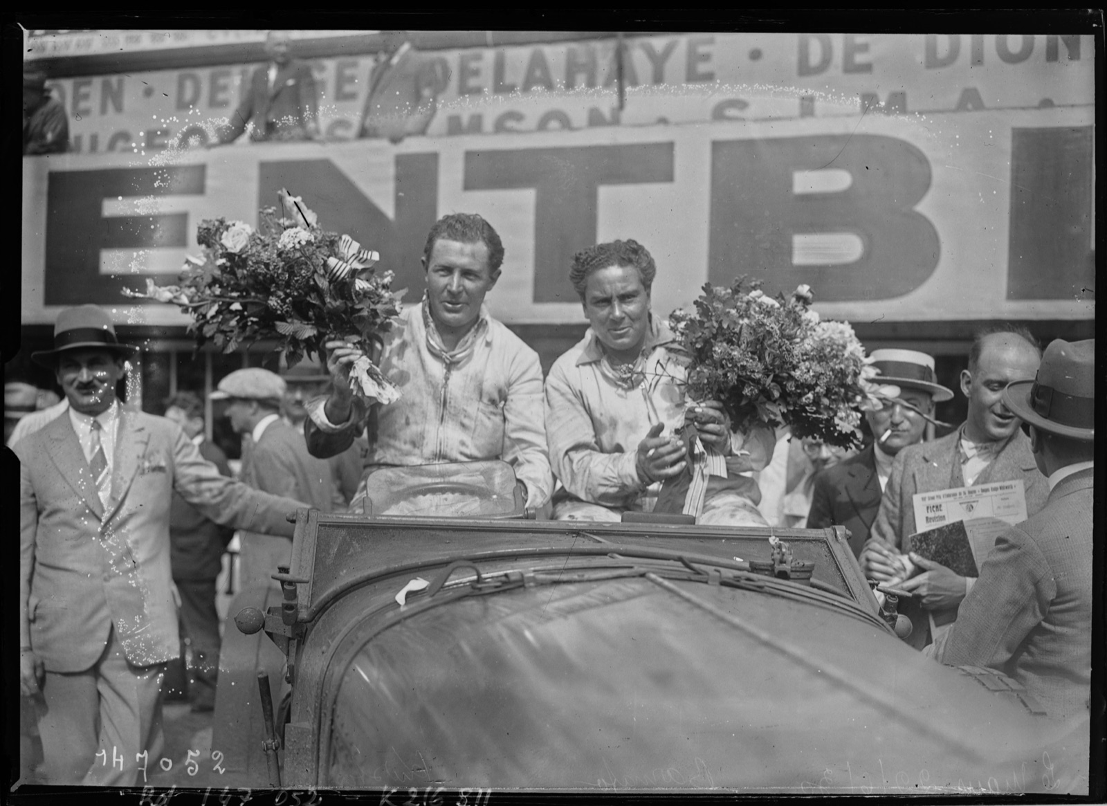 Glen Kidston and Woolf Barnato in oil-stained overalls holding bouquets in their Bentley in front of a large Bentley banner after winning Le Mans in 1930