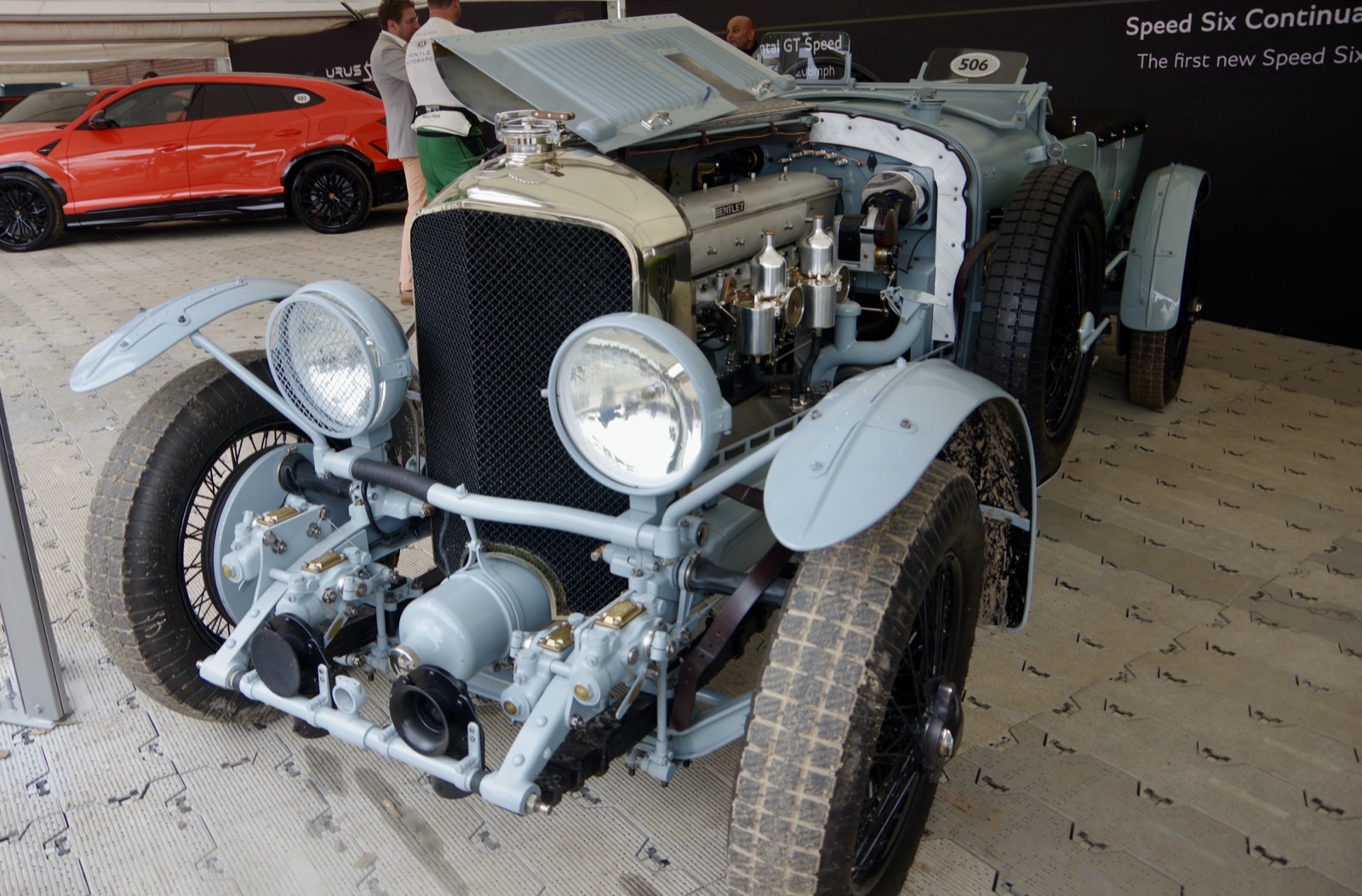 A pale blue-grey Bentley Speed Six continuation car with its bonnet open showing the six-cylinder engine and twin carburettors, on a stand at Goodwood