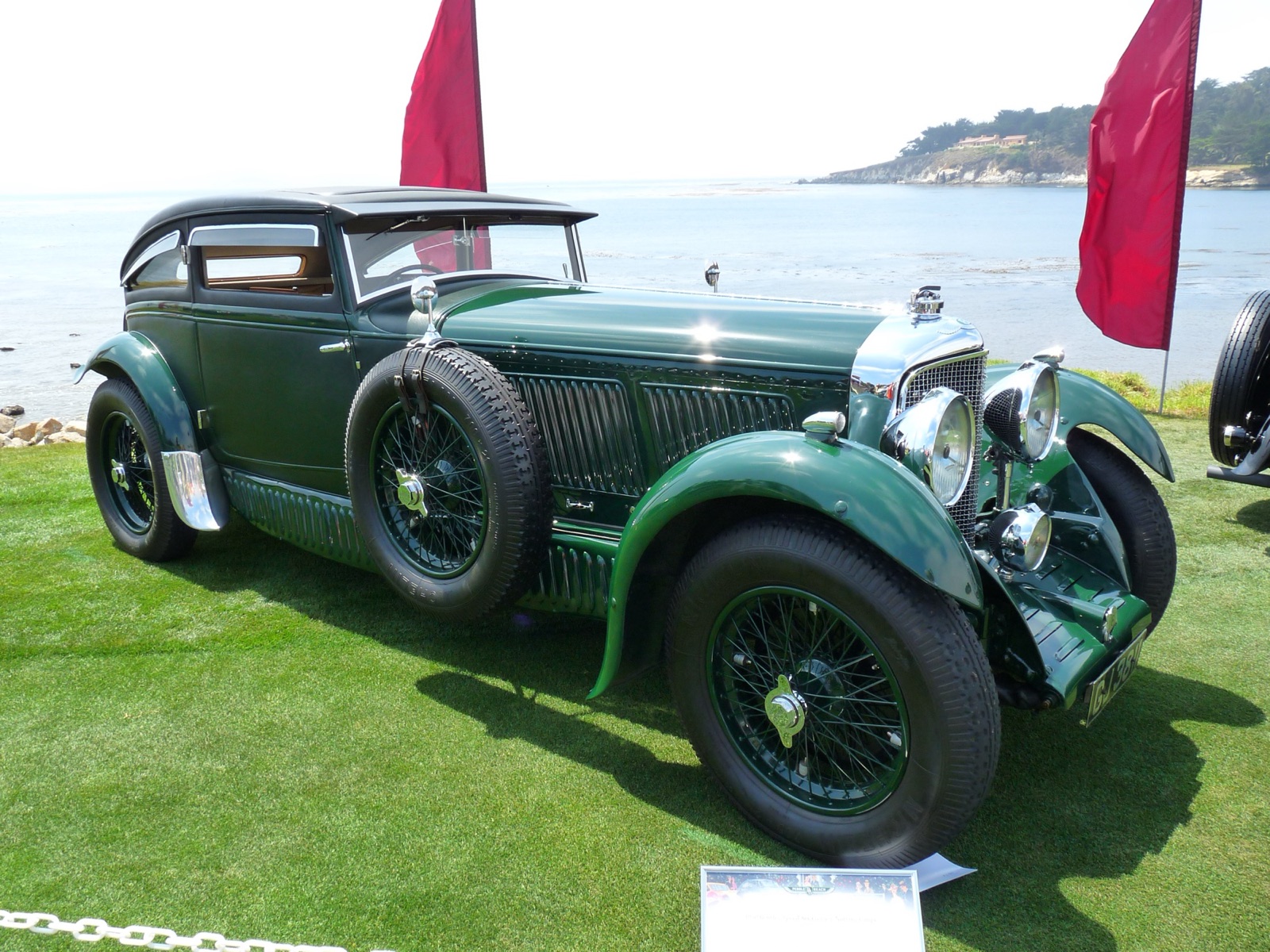 A dark green Bentley Speed Six coupé with a low roof, side-mounted spare wheel and long louvred bonnet on the lawn at Pebble Beach, with the sea behind