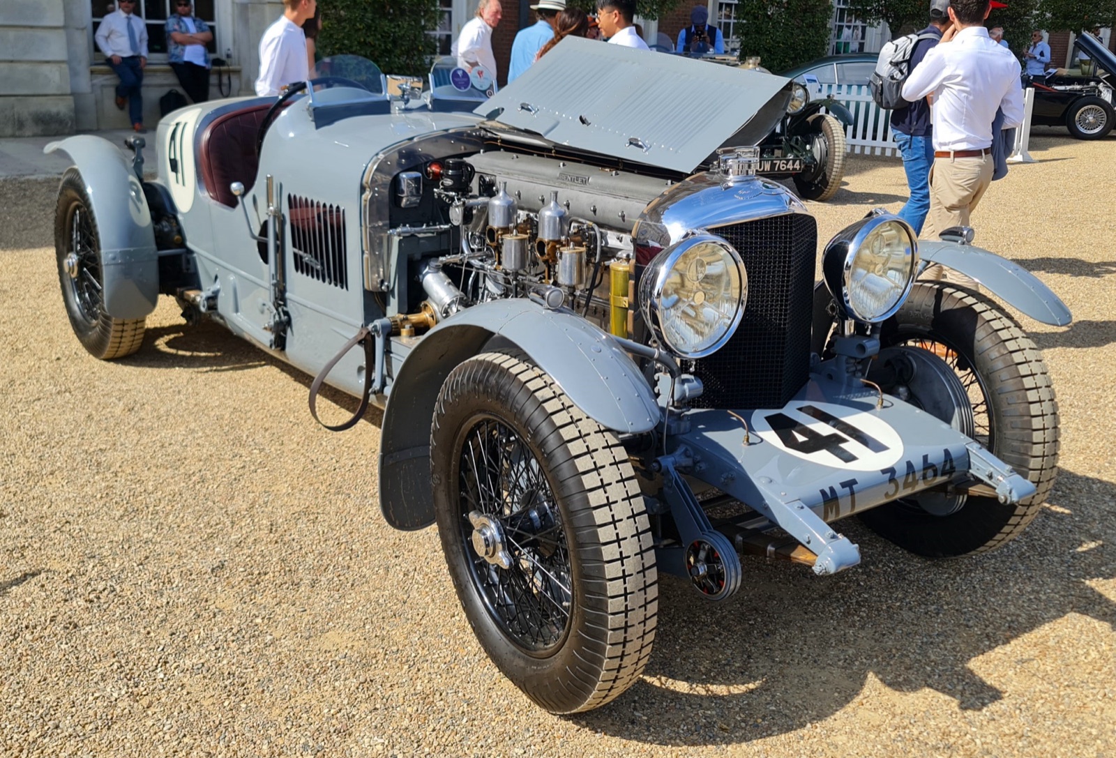 A grey Bentley two-seater racing car with its bonnet open showing a six-cylinder engine, racing number 41 and registration MT 3464, on gravel at a concours