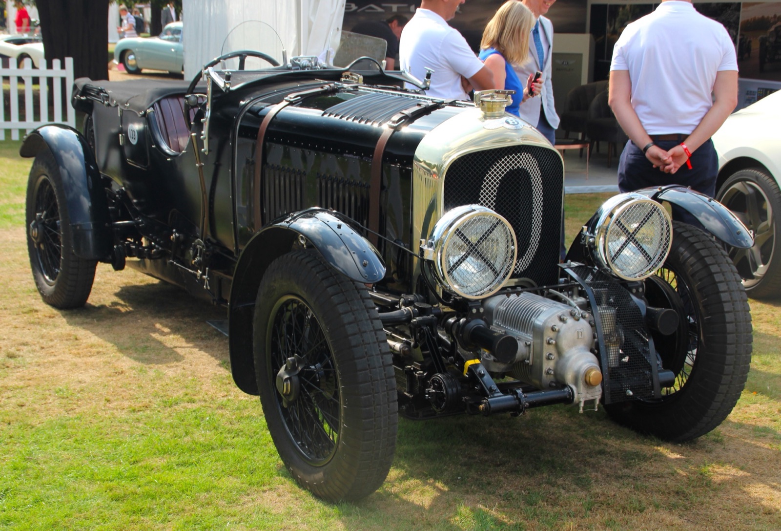 A black Blower Bentley continuation car with a polished radiator shell, the number 0 on its radiator mesh, the supercharger in front and leather straps across the louvred bonnet, parked on grass