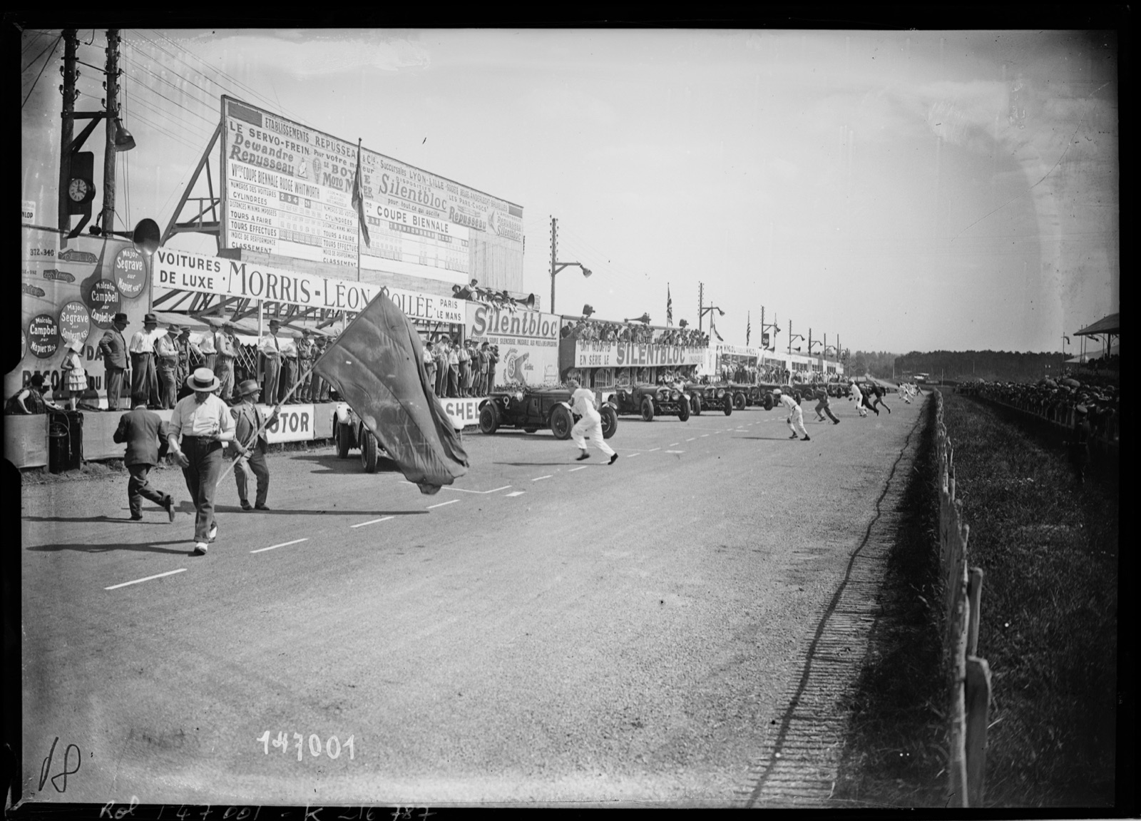 Le Mans start line in 1930: a man waves a large flag in the foreground while drivers in white overalls run across the road to cars parked at an angle along the pits, Bentley number 9 just beyond the flag and numbers 3 and 4 behind it