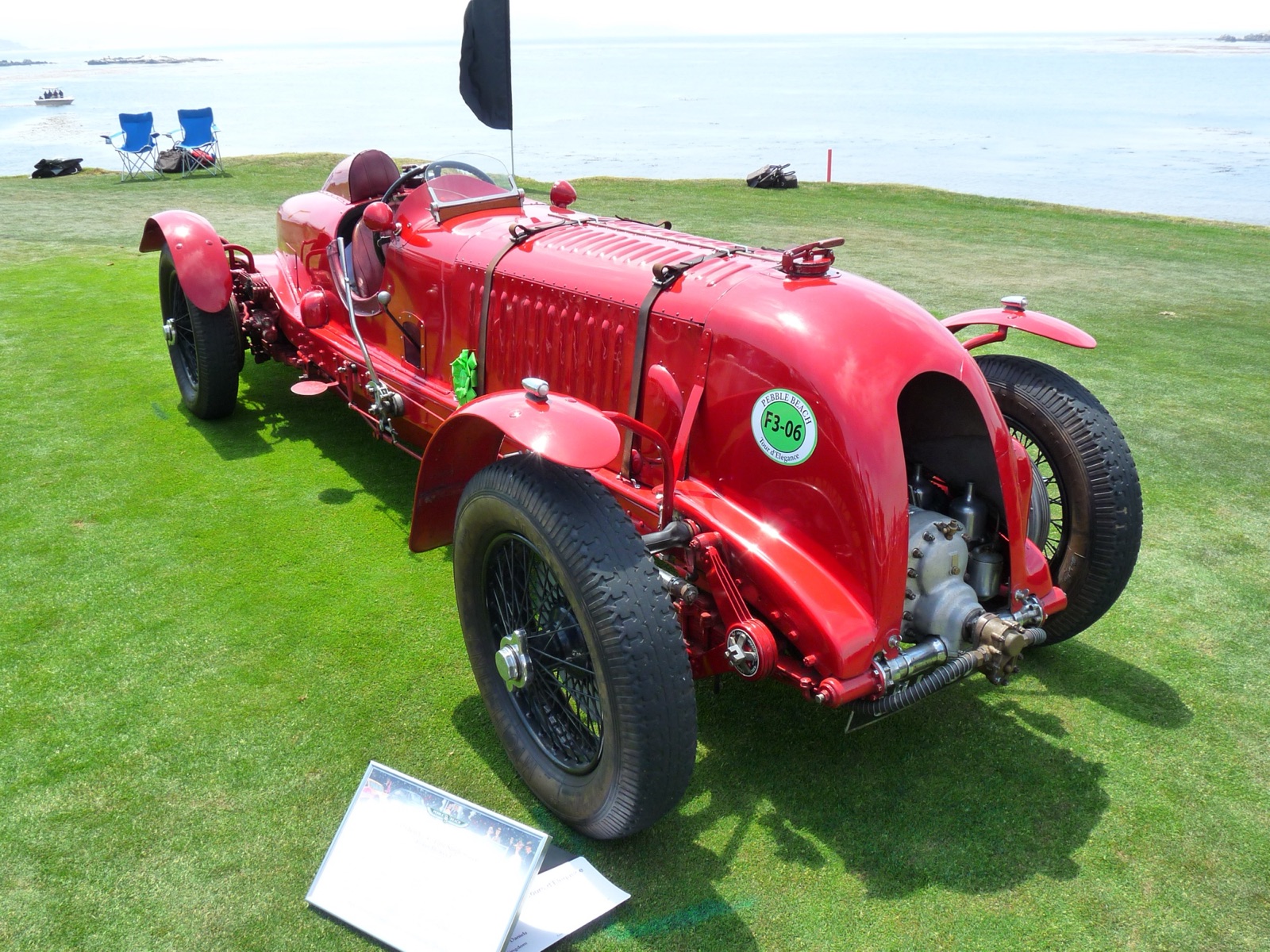 A long red single-seat Bentley racing car on grass by the sea, with cycle wings, a louvred bonnet held by a leather strap and the supercharger exposed at the front below the cowled radiator