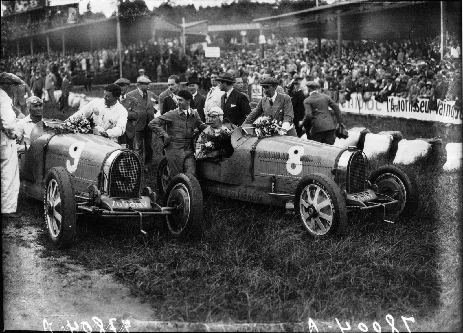 Two Bugatti Grand Prix cars, numbers 9 and 8, decked with flowers in front of a crowded grandstand at Spa