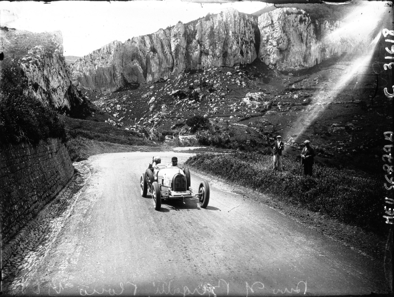Albert Divo driving a Bugatti Type 35 along a mountain road below limestone cliffs in Sicily, two spectators at the roadside