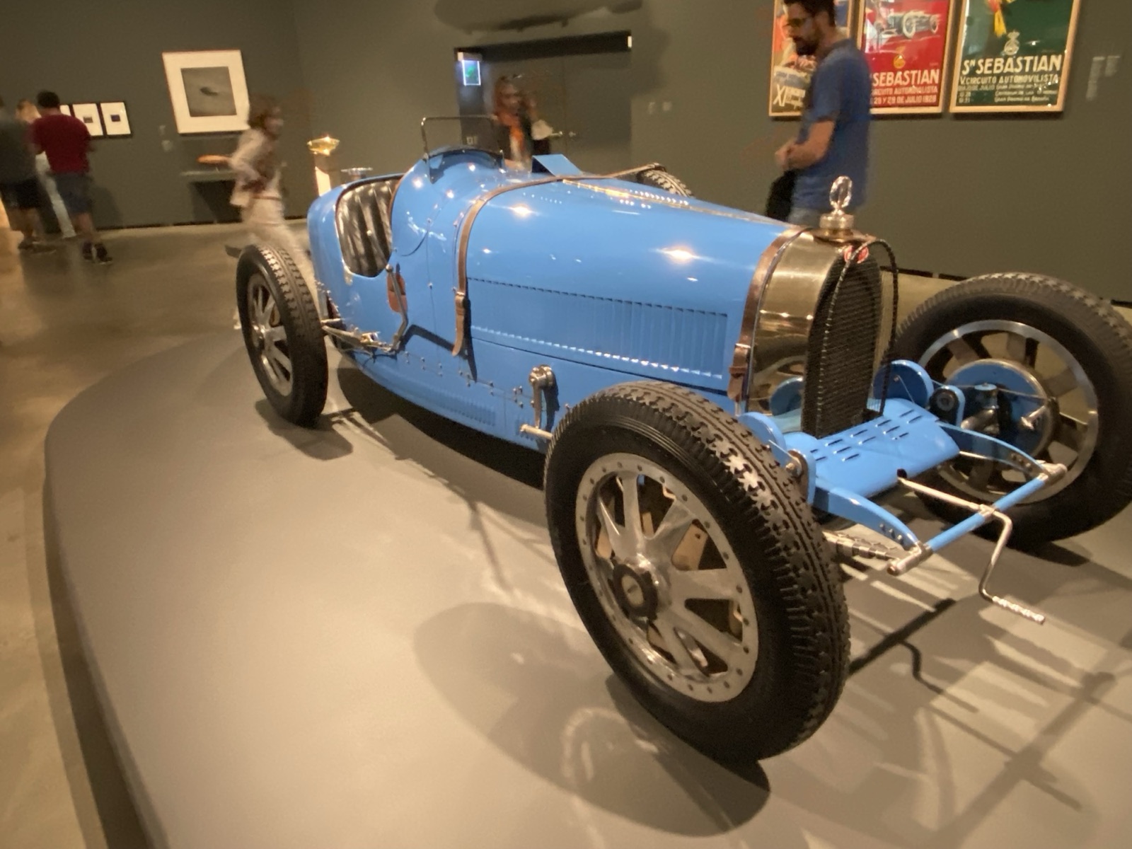 Pale blue Bugatti Type 35 with cast wheels on display in a museum gallery, racing posters on the wall behind