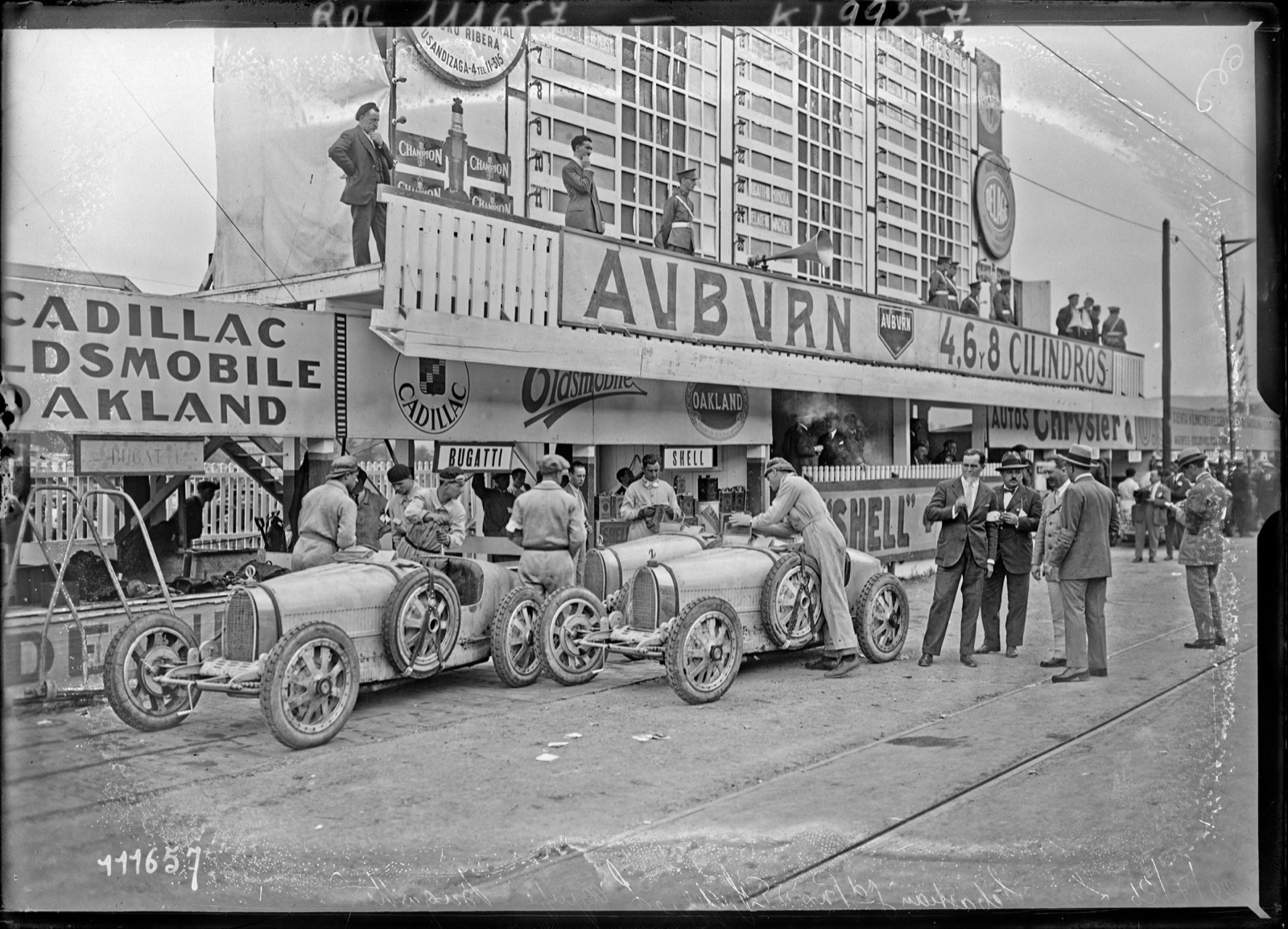 Three Bugatti Grand Prix cars with mechanics in front of the advertising-covered pits at San Sebastián