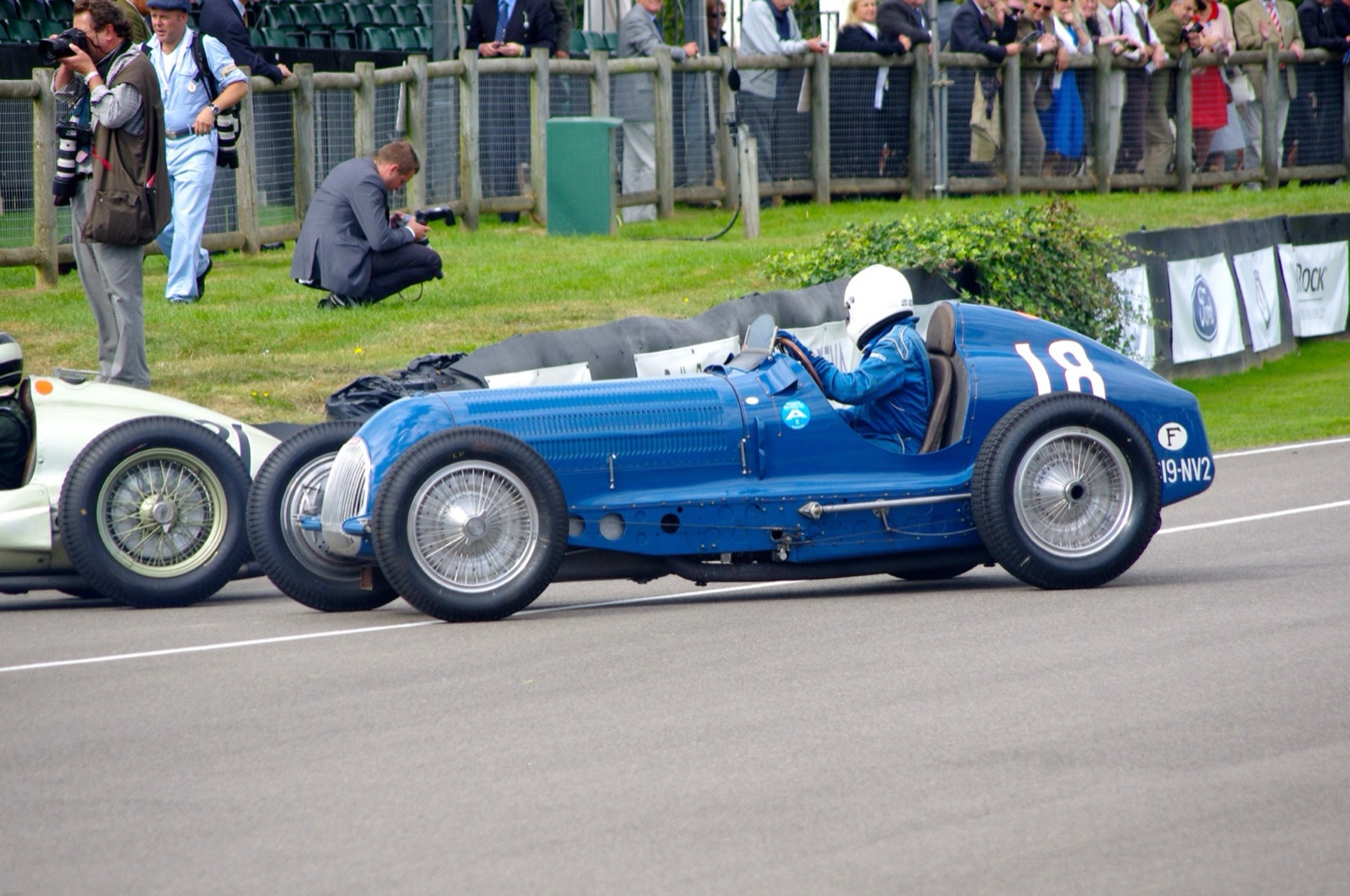 A blue single-seat Bugatti Type 59/50B III with a cowled radiator and number 18, driven past photographers on a circuit