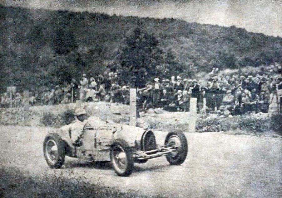 Period photograph of René Dreyfus driving a Bugatti Type 59 through a bend past a crowd behind a fence, with a wooded hill behind