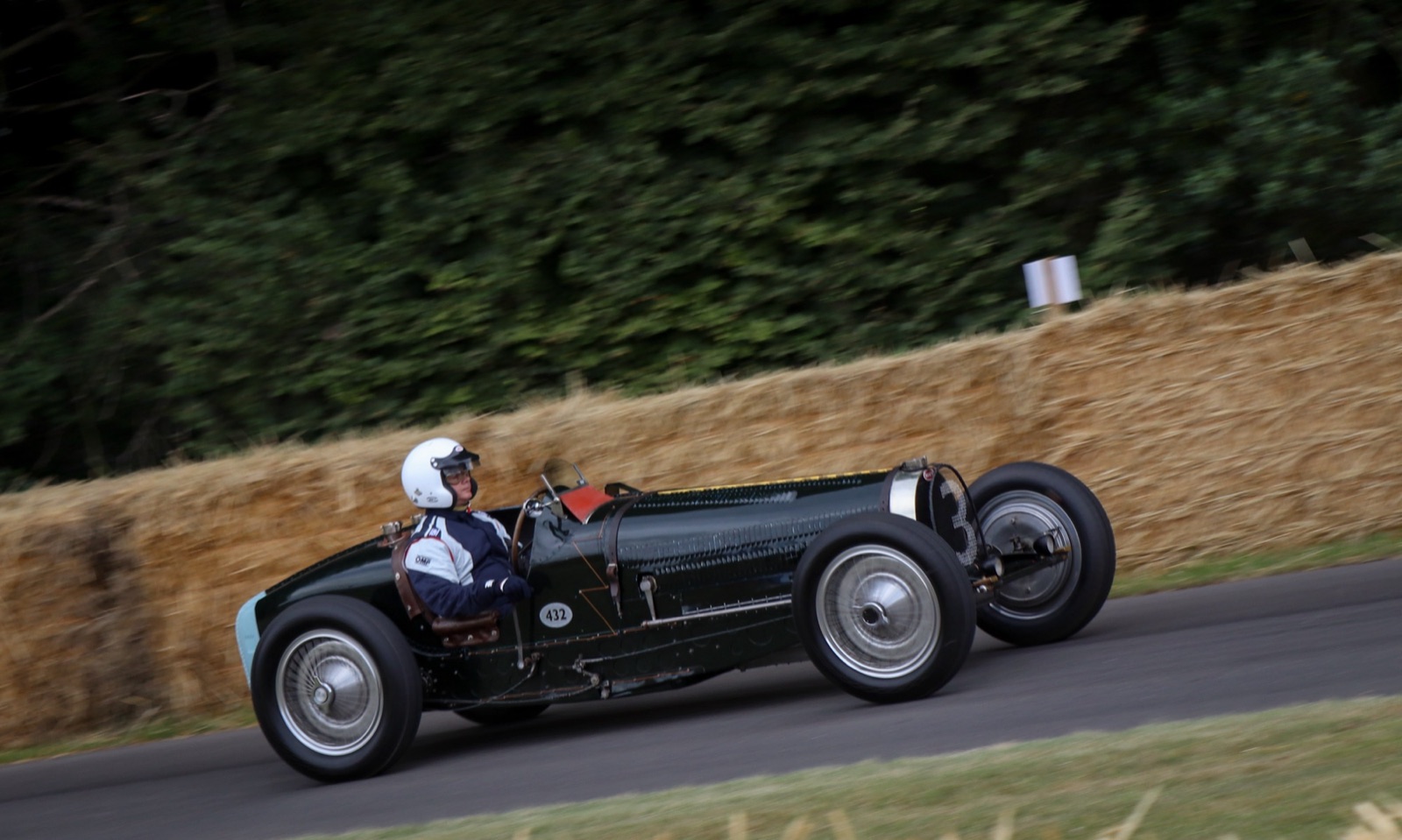 A dark green Bugatti Type 59 with piano-wire wheels driven past straw bales by a driver in a white helmet