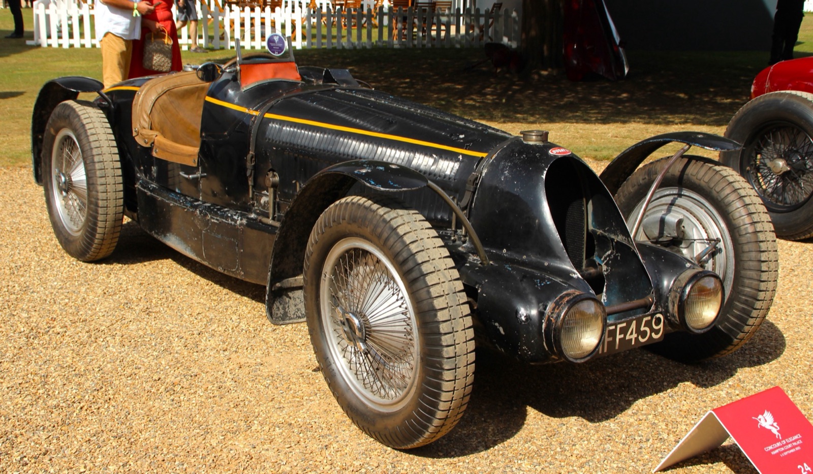The black Bugatti Type 59 Sports with a yellow stripe, cycle wings, headlamps and worn paint, on gravel at a concours