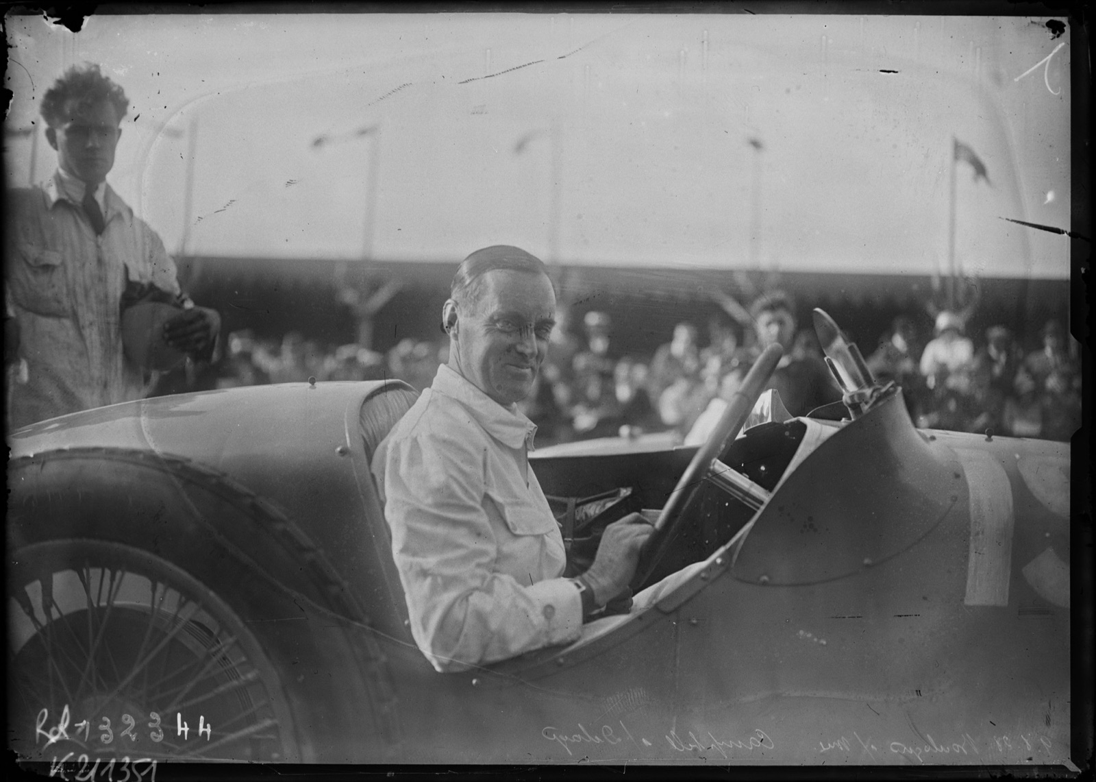 Malcolm Campbell in a white overall smiling from the cockpit of his Delage, a young man standing behind the car