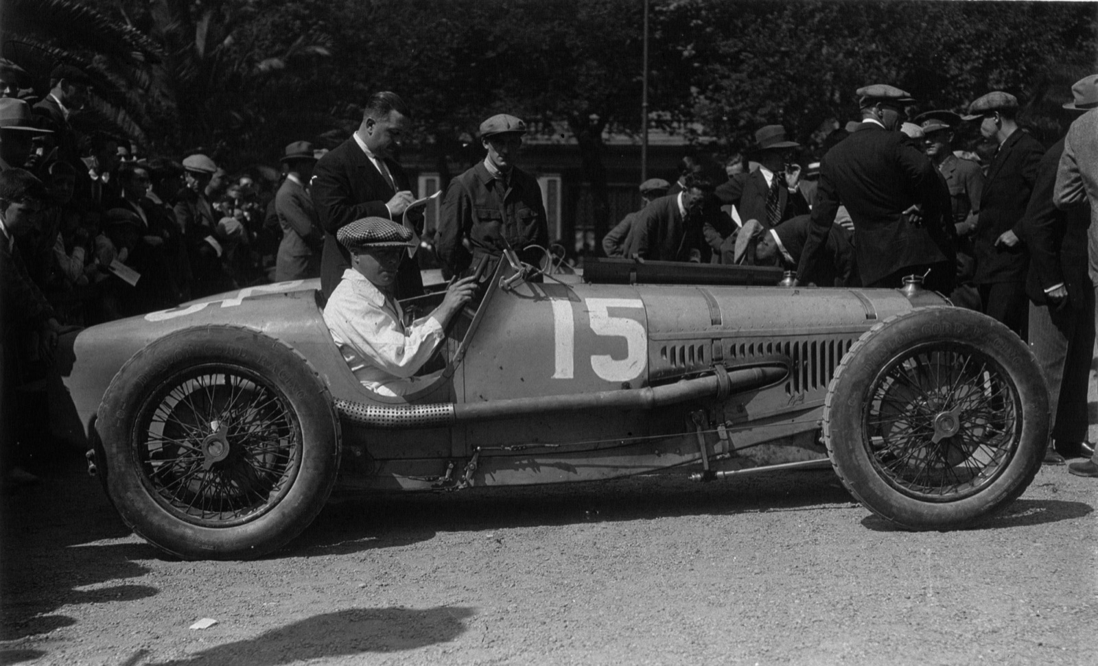 A driver in a white overall seated in a Delage 15 S8 numbered 15, the exhaust pipe running along the right side beside the cockpit, crowd behind
